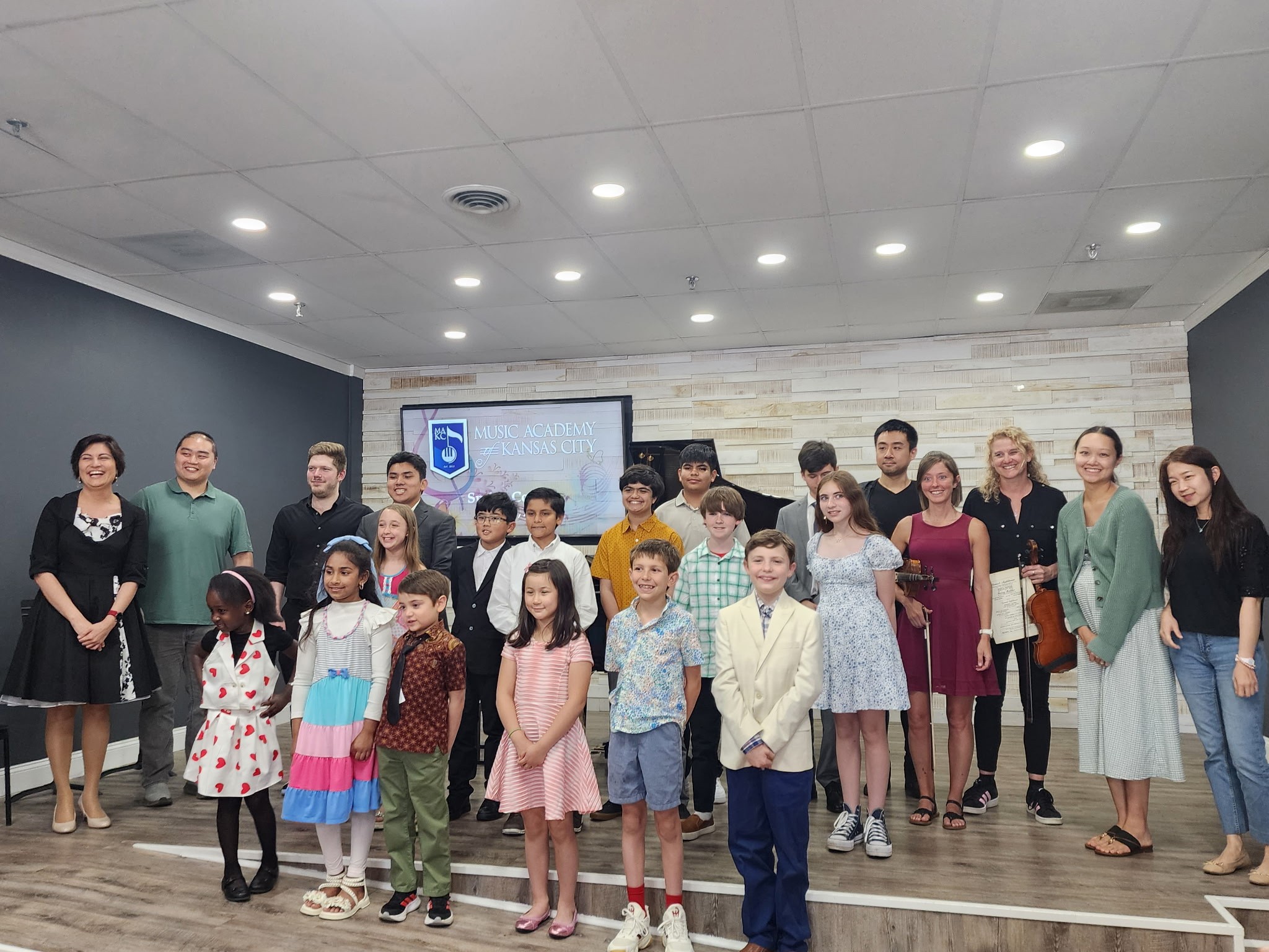 Group of children and adults standing indoors, some smiling, in a room with a white brick wall and ceiling lights.