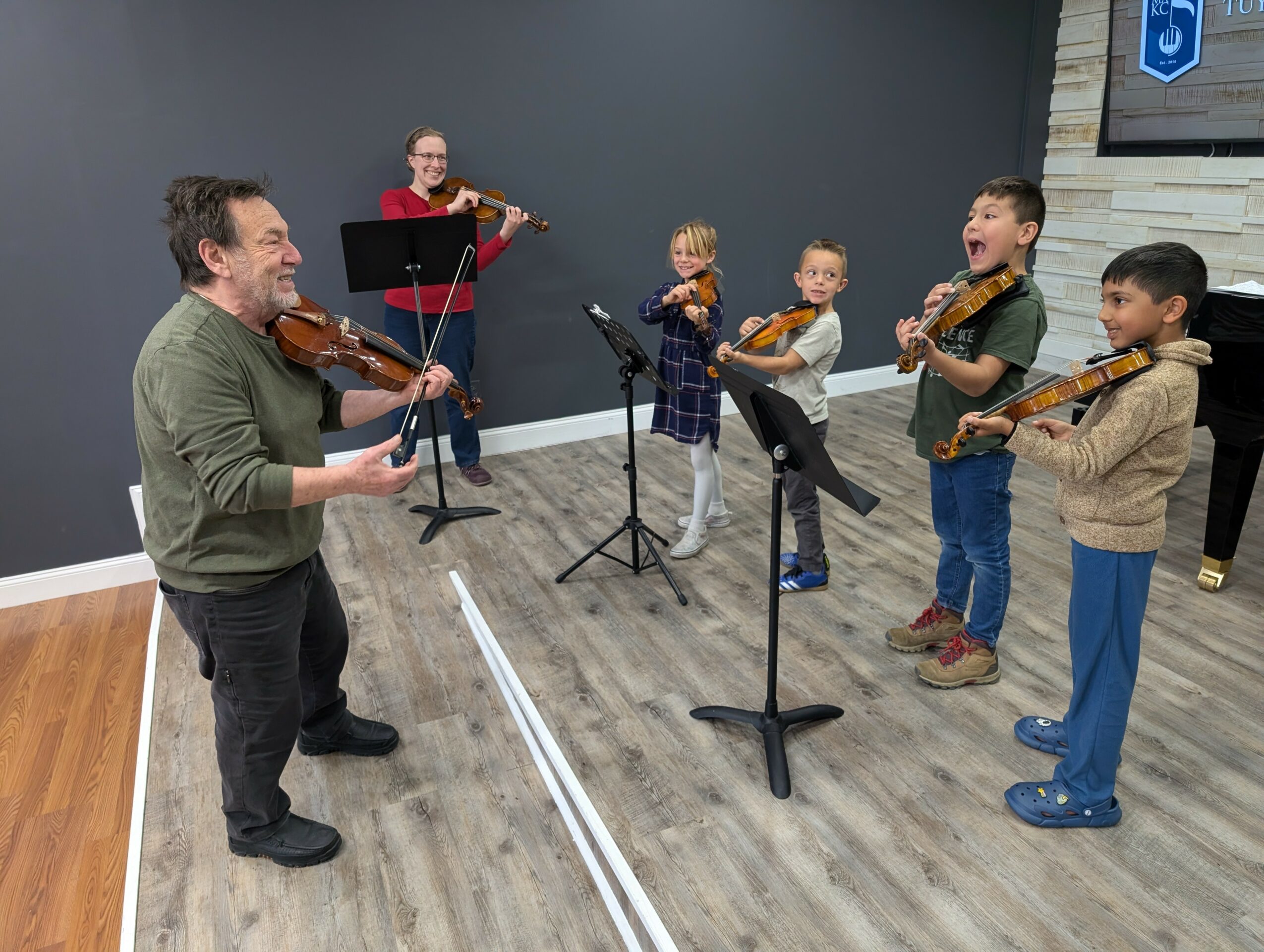 Adult and children playing violins in a music class, standing in a room with dark gray wall and wooden floor.