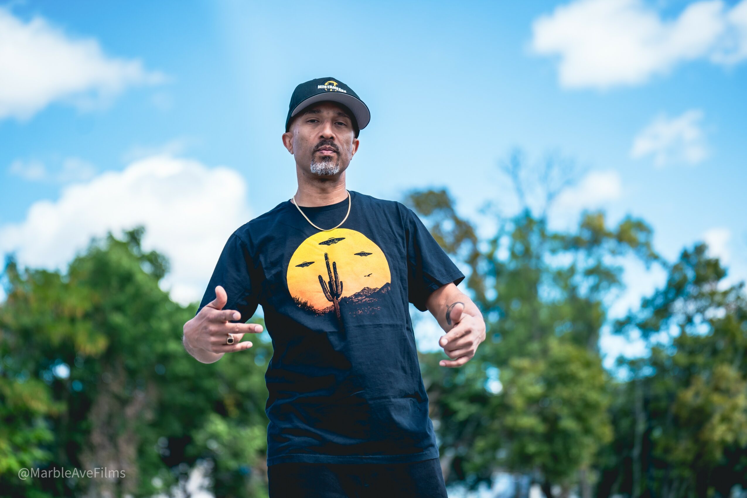 Man wearing a black T-shirt with a desert sunset graphic, standing outdoors with trees and blue sky behind him.