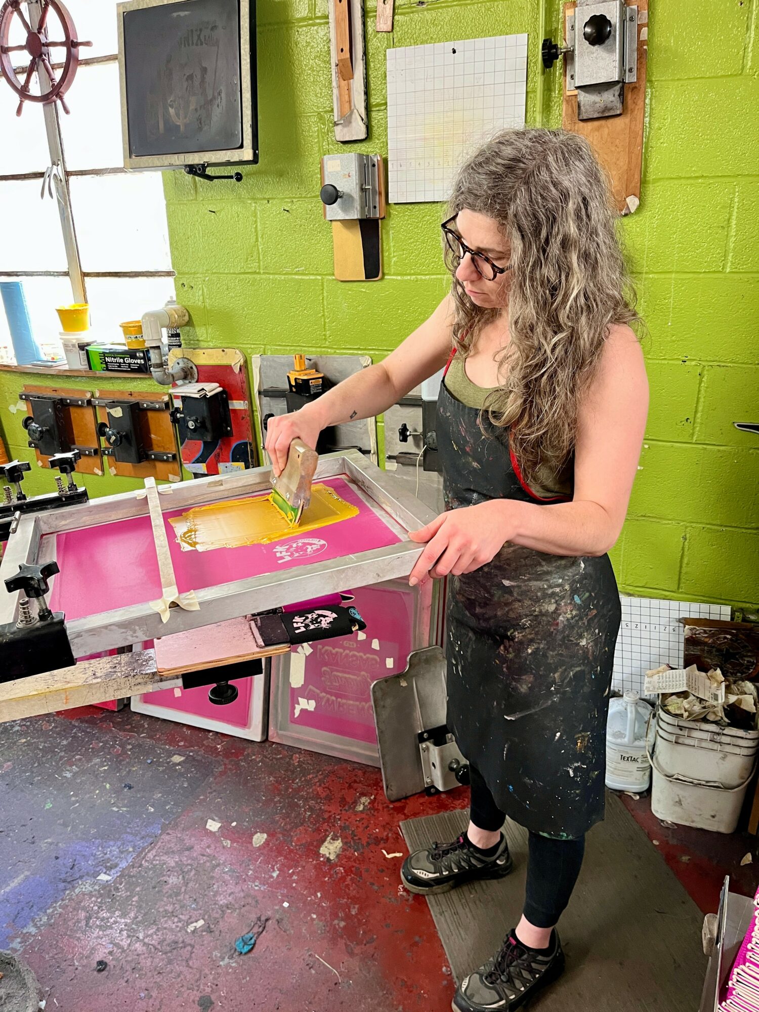 Girl with curly hair and glasses working at a screen printing press in a workshop with green walls.