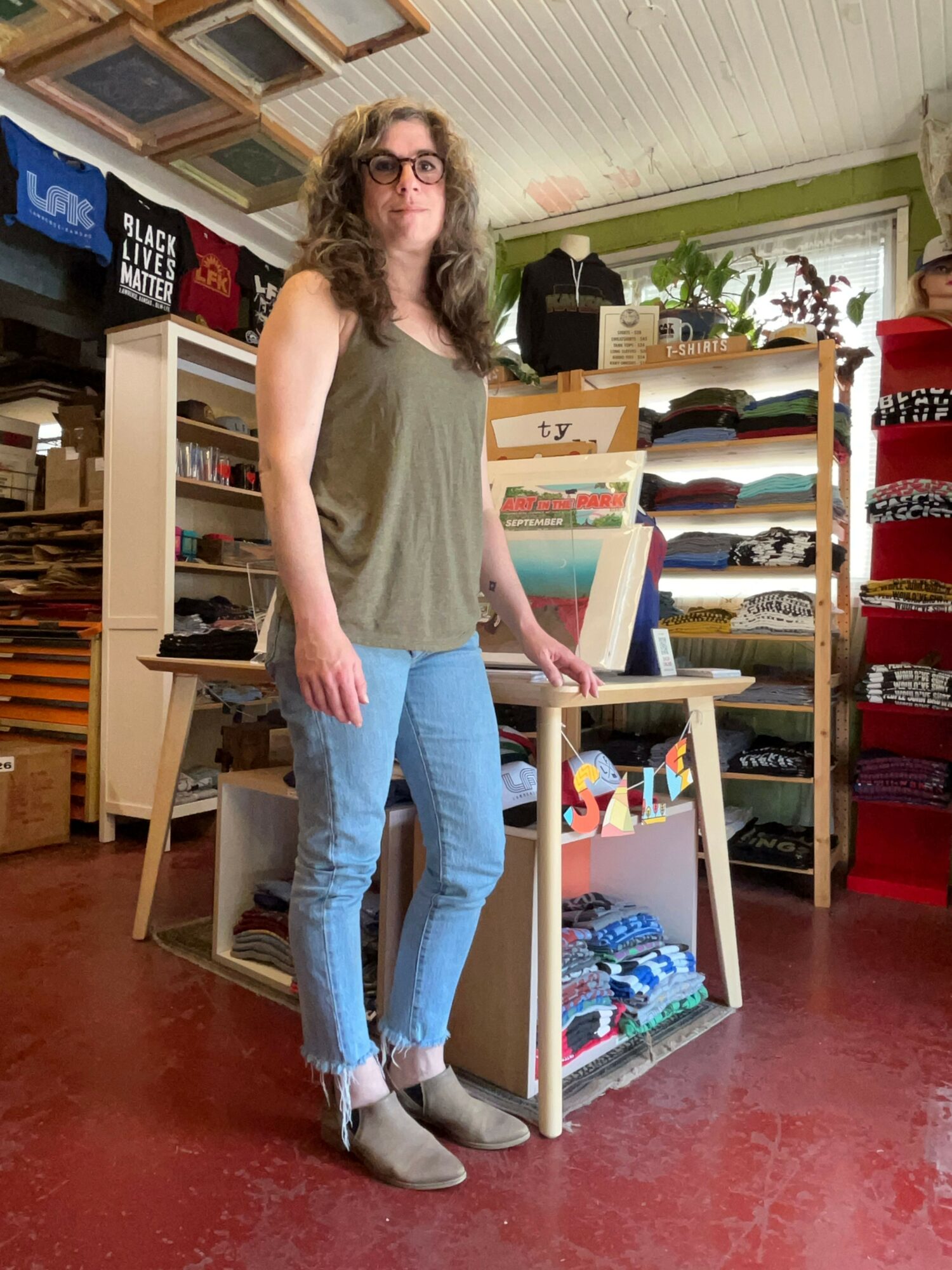 Woman standing next to a display table with books in a bookstore, holding a book. Shelves with books are in the background.