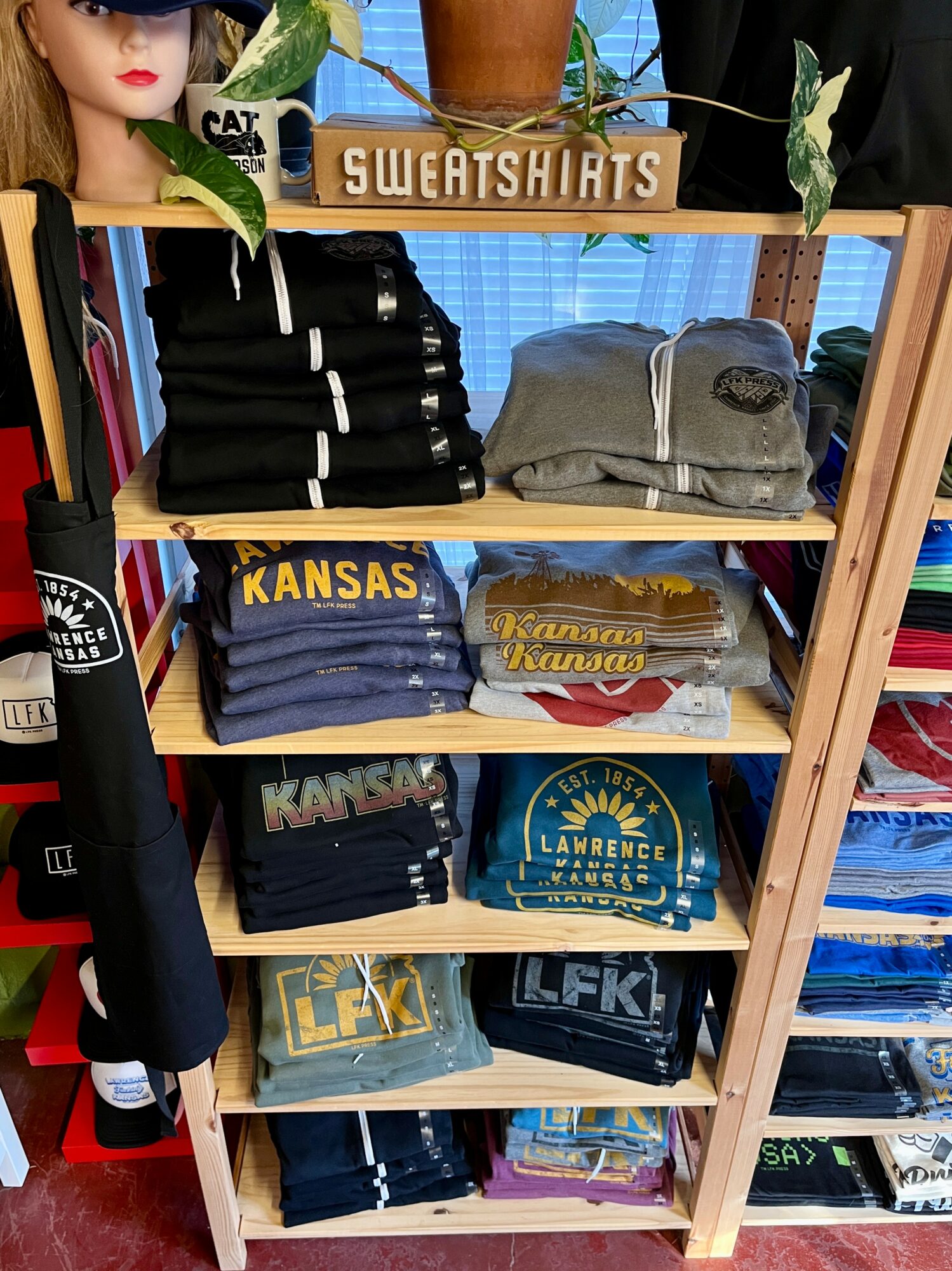 Shelf displaying folded sweatshirts and T-shirts with Kansas and LFK logos, with a mannequin head and plants on top.