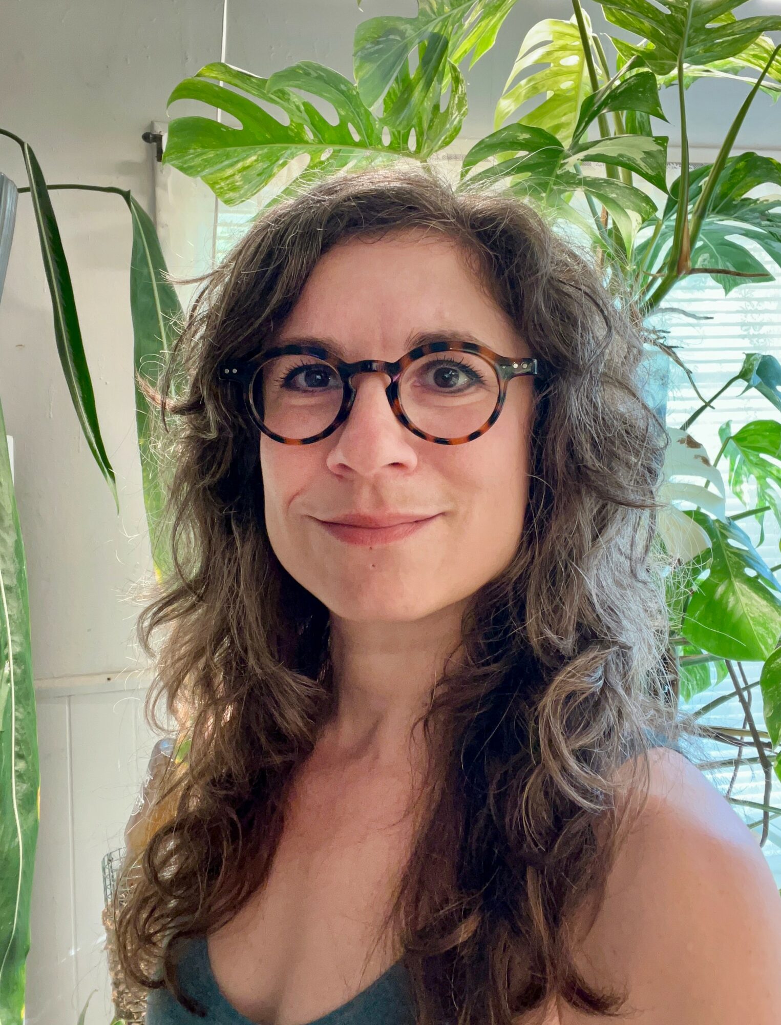 Woman with curly hair and glasses smiling indoors with large green plants behind her.
