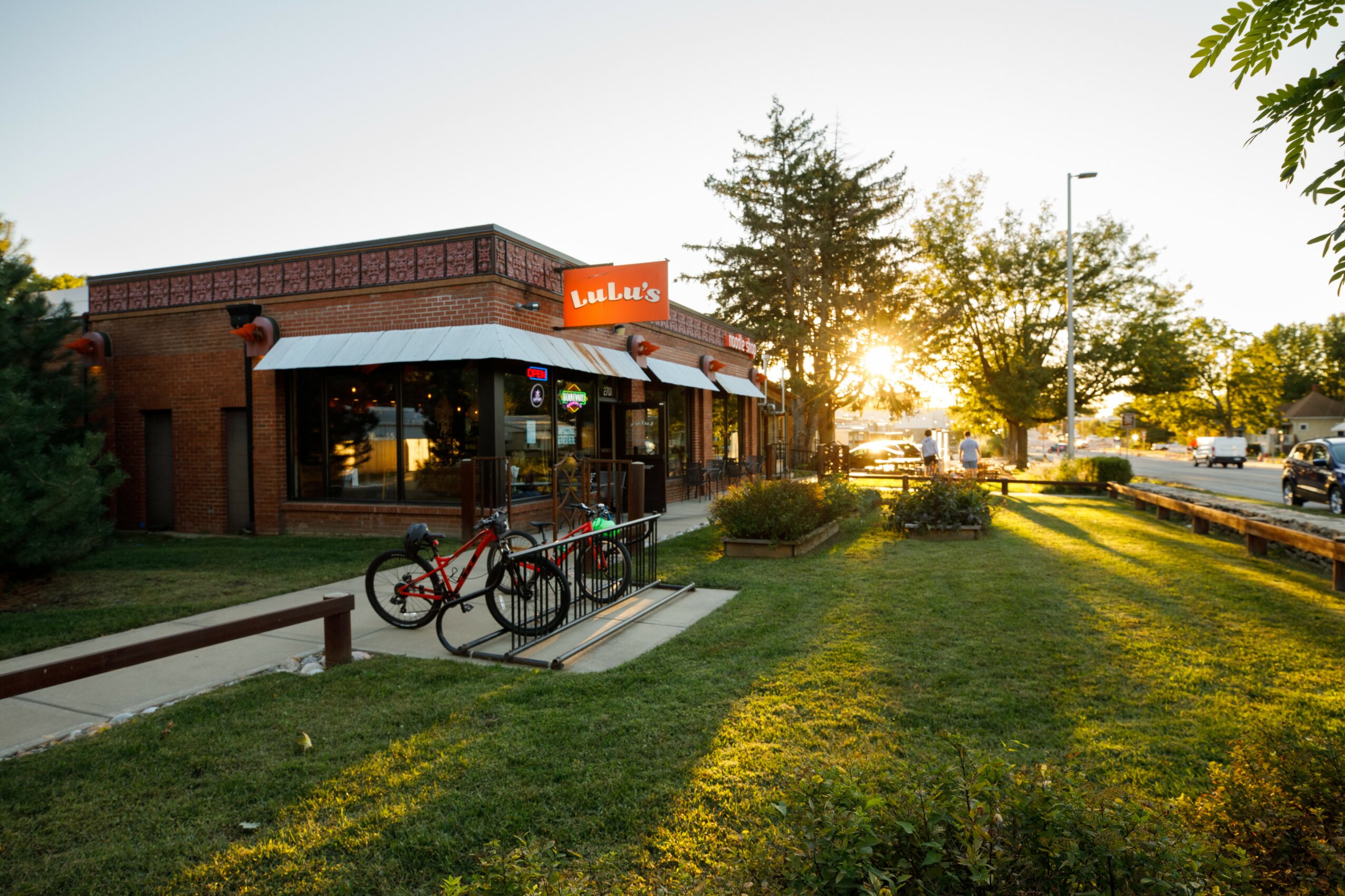 A brick building with a white awning and bicycles outside, sun setting behind trees, casting long shadows on the grass.
