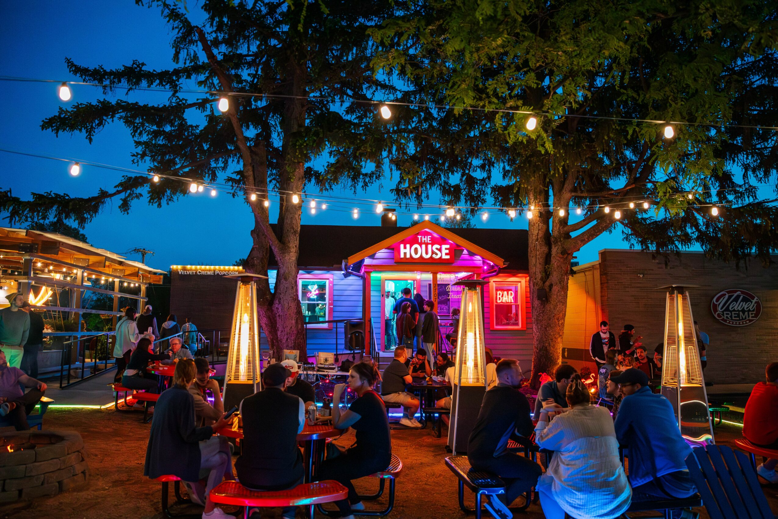 Night scene with people sitting at tables outdoors, illuminated by string lights and a colorful house-shaped stage, trees in background.