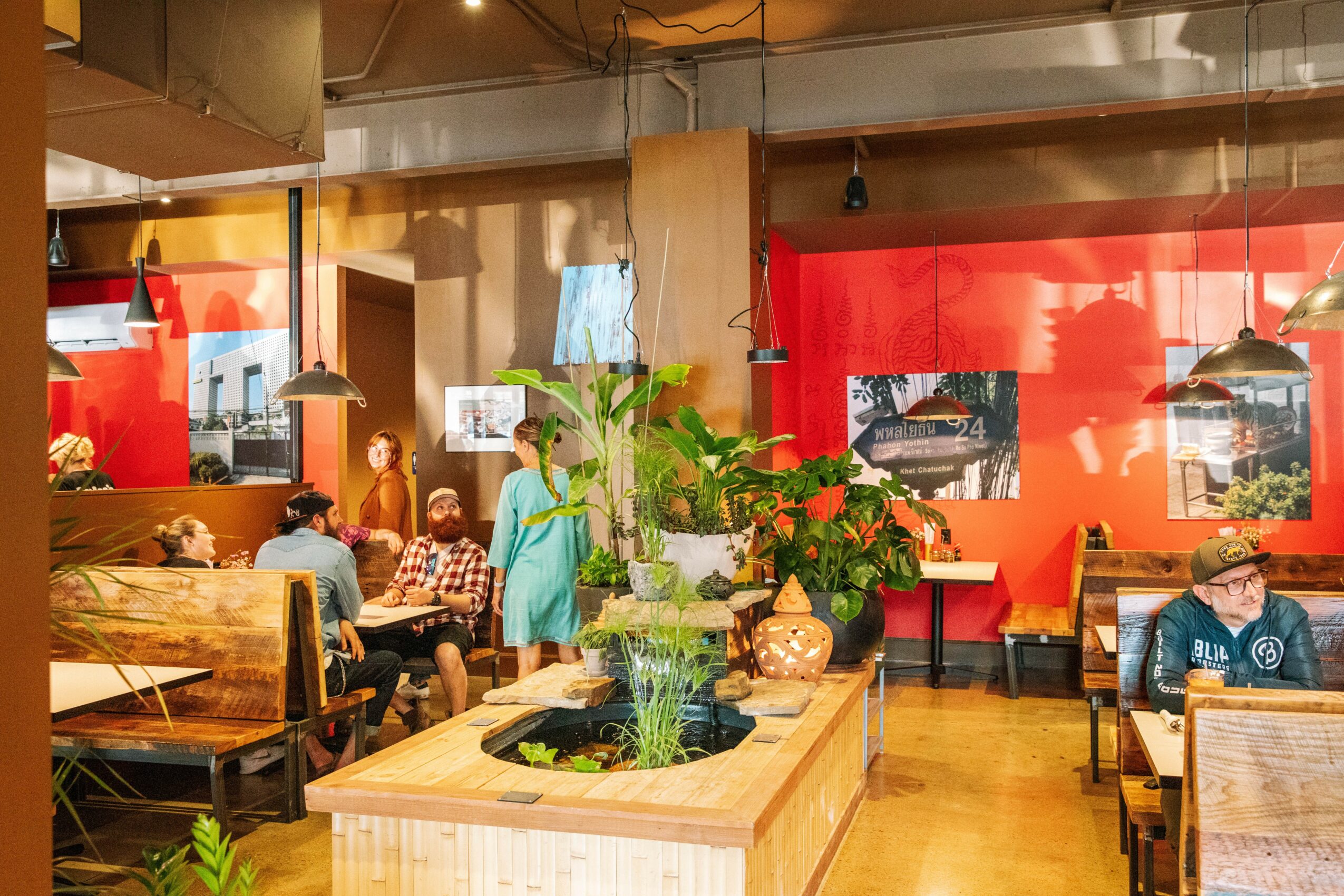 People dining in a restaurant with red and orange walls, plants, and wooden furniture, some people sitting and talking.