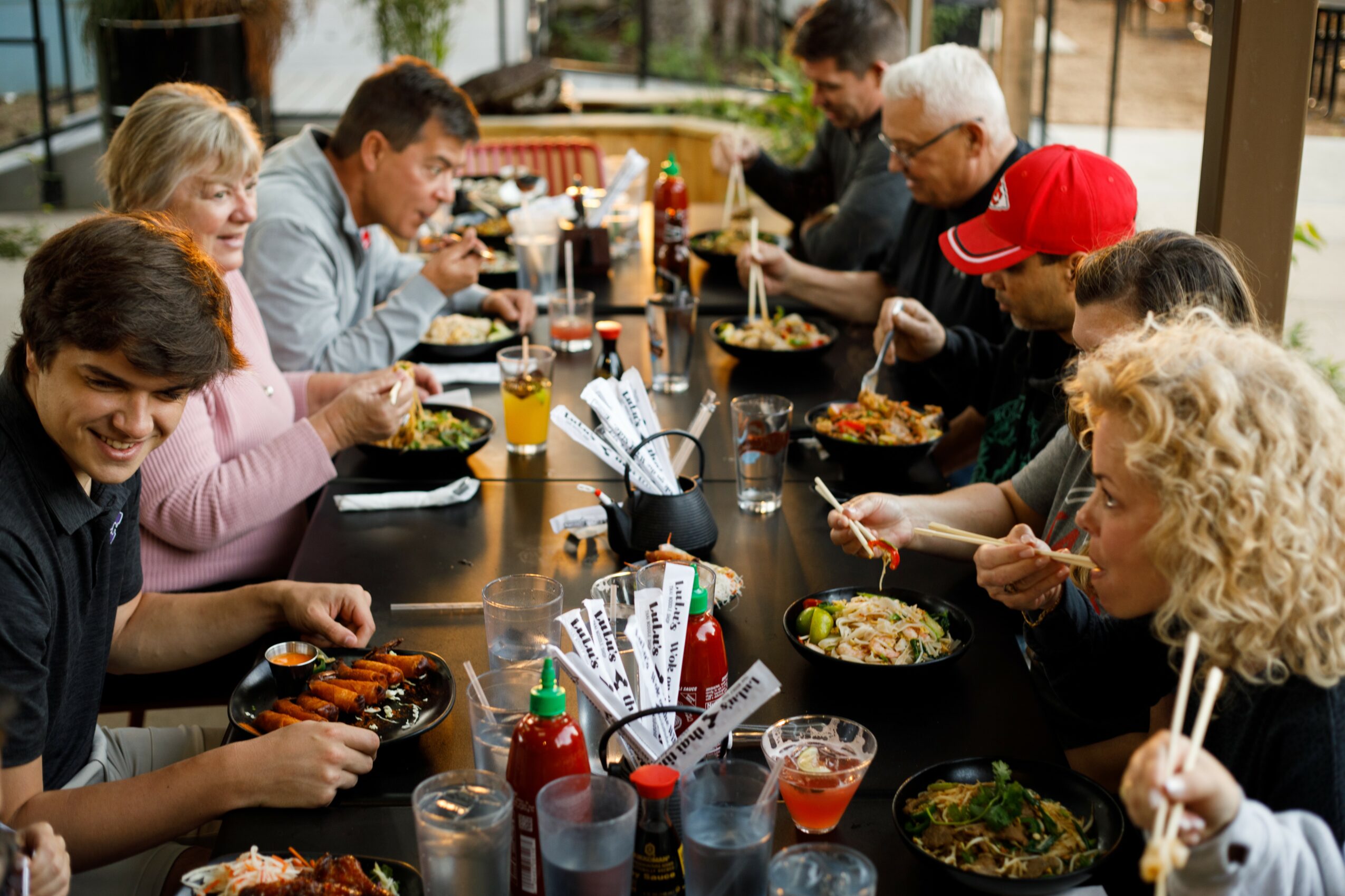 Group of people dining at a long table with food and drinks, indoors with large windows, smiling and eating.