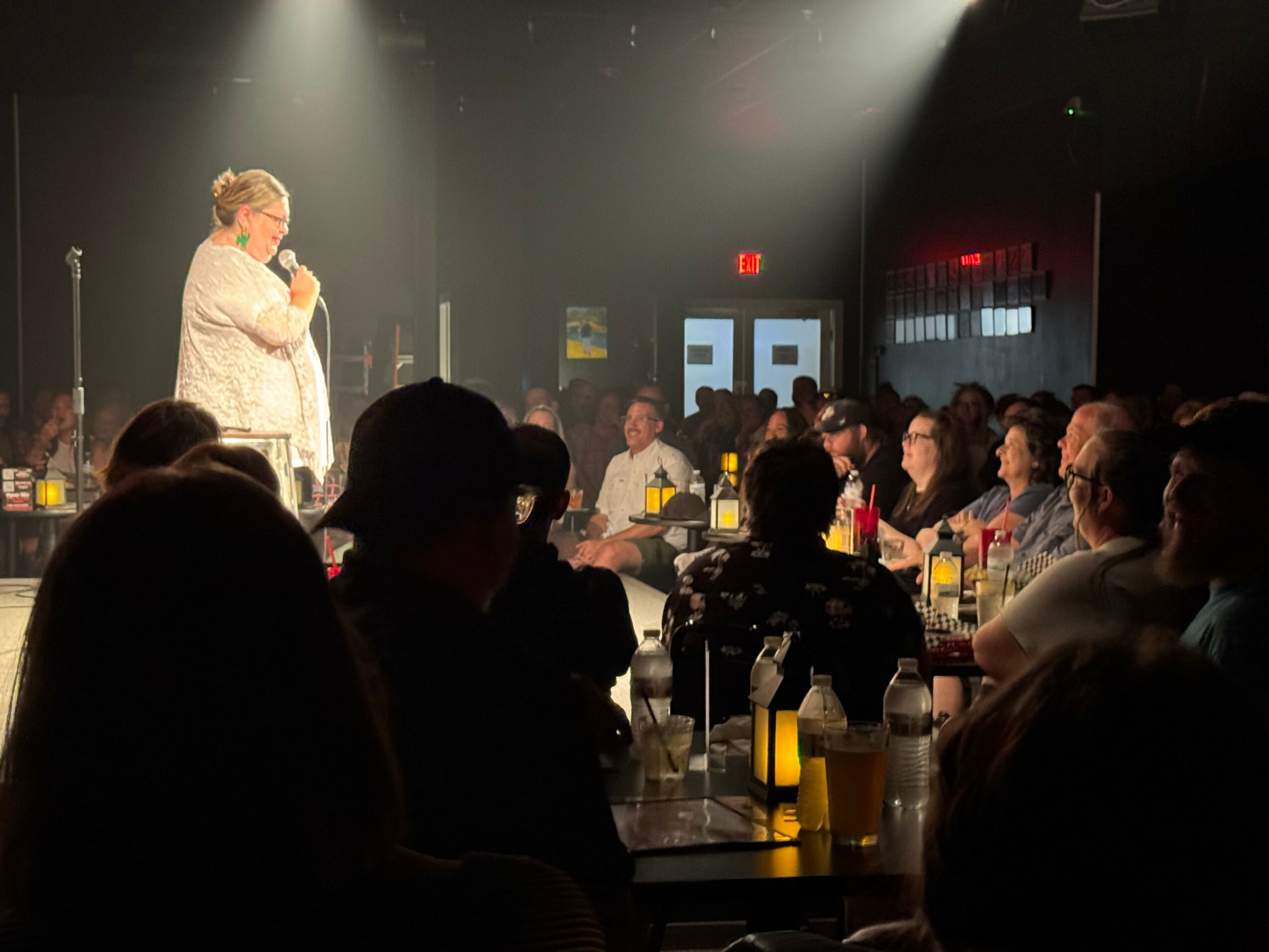 A woman stands on stage speaking into a microphone, audience seated at tables in a dimly lit venue.