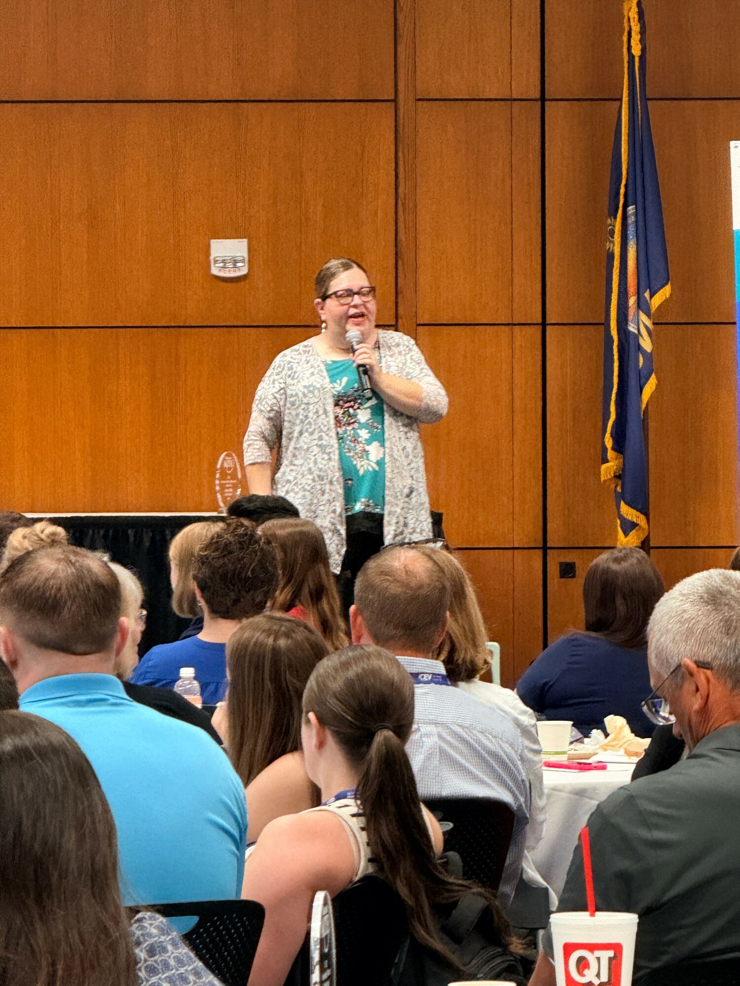 A woman speaking to a group of people in a room with wooden walls, some seated and some standing.