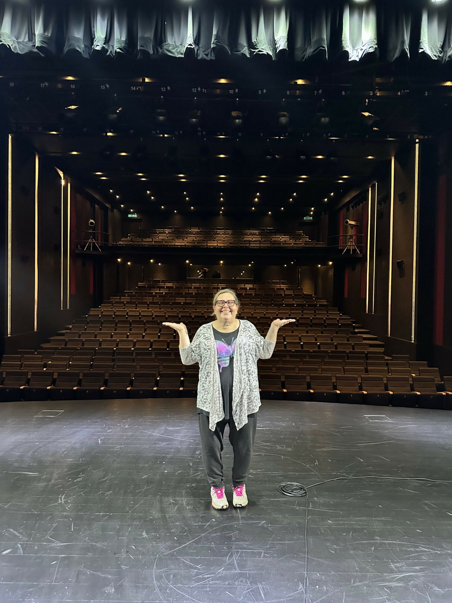 Person standing in an empty theater with rows of seats and a stage, waving at the camera.