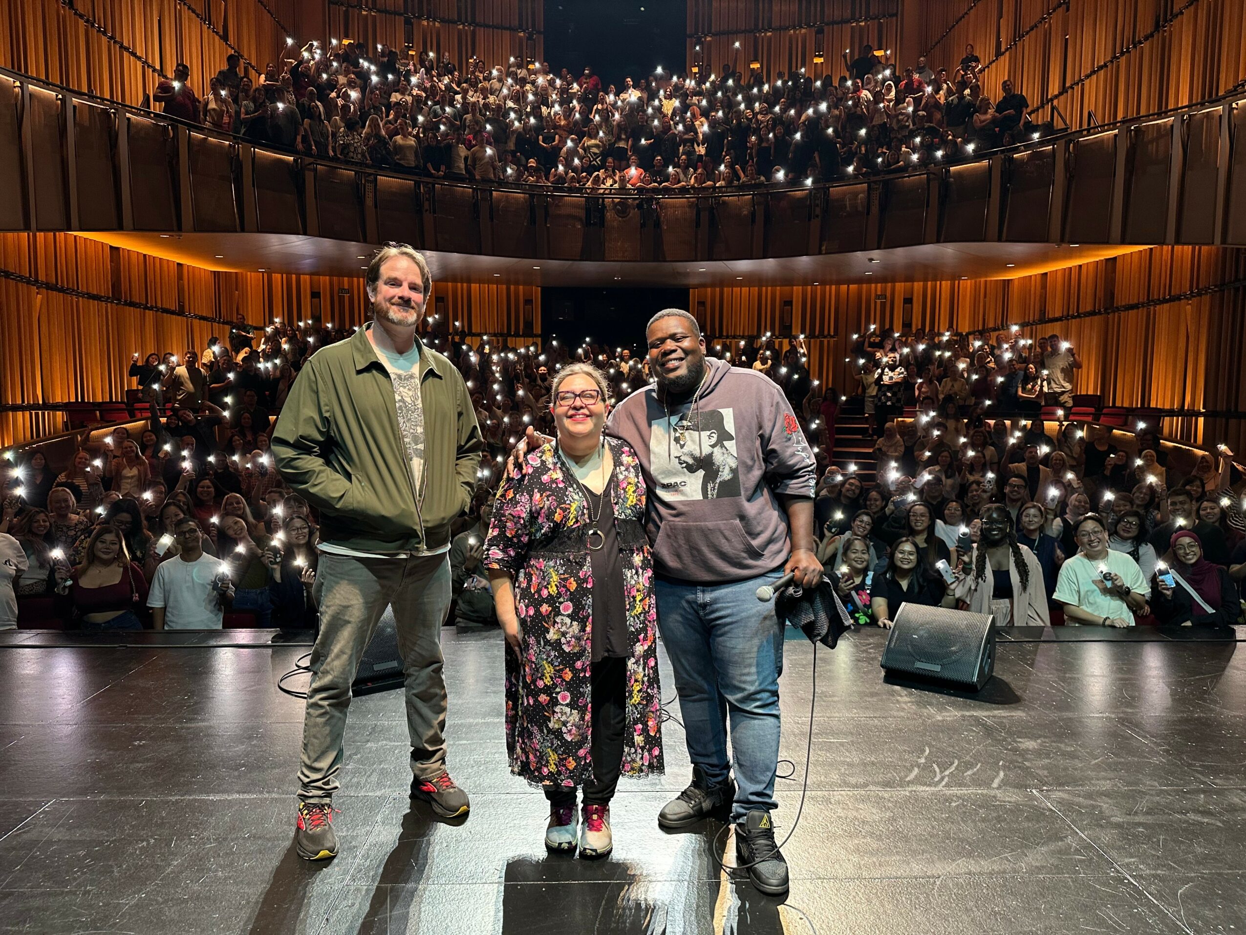 Three people standing on stage in front of an audience in a concert hall, with audience members holding lights. Word count: 20