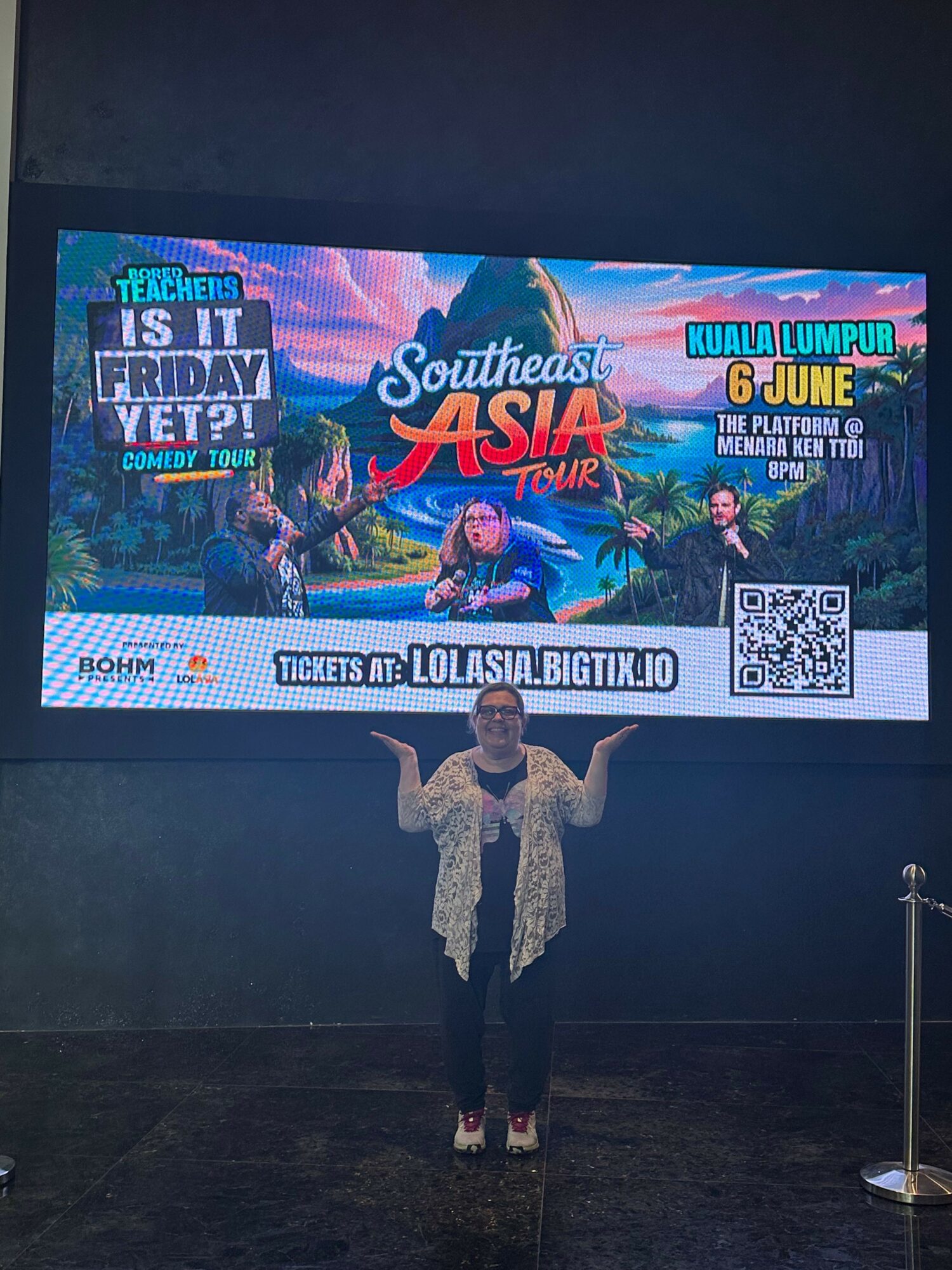Person standing in front of a colorful Southest Asia-themed event poster, holding a phone, with a dark background.