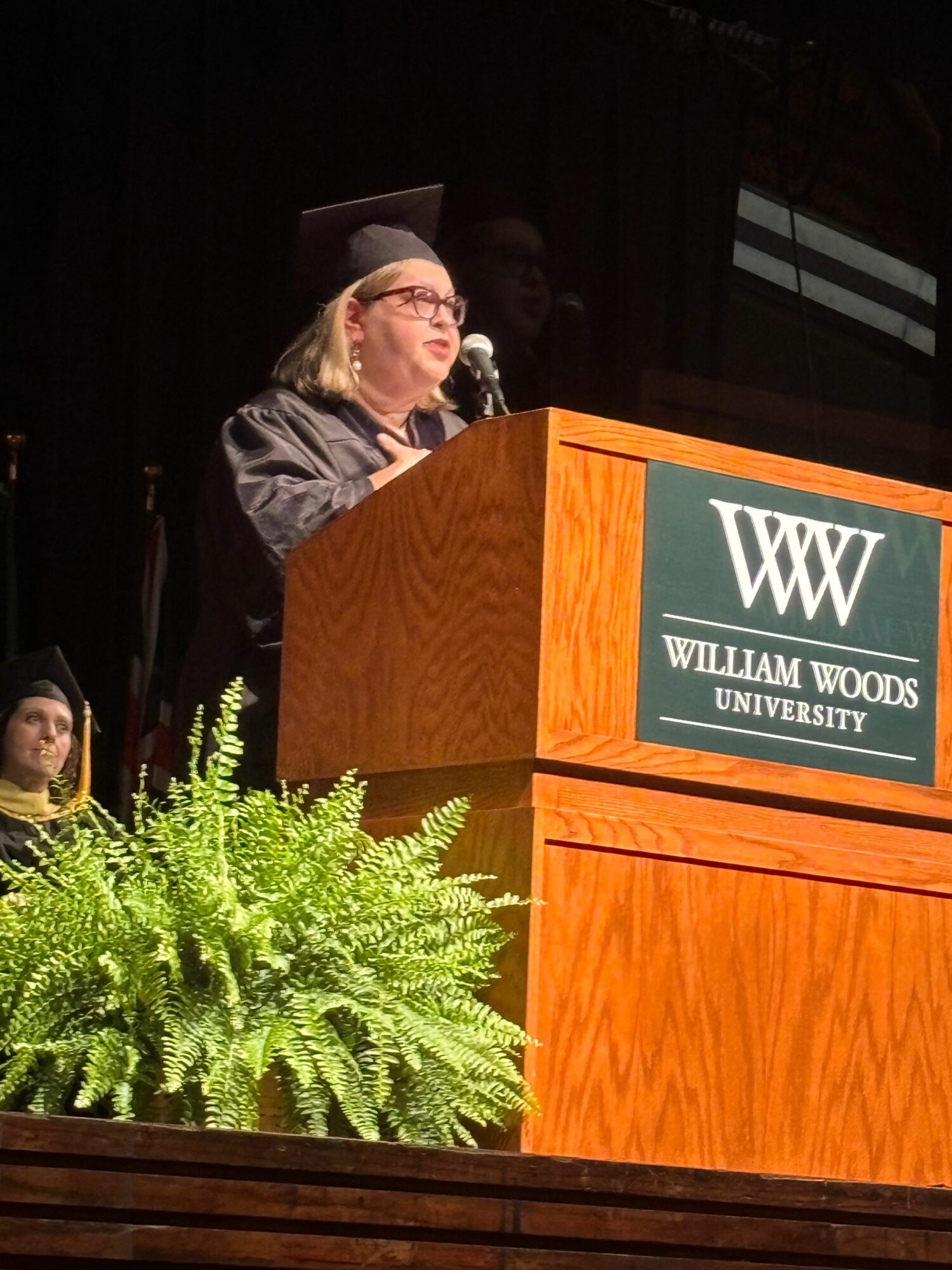 Person speaking at a podium with William Woods University logo, green plant in foreground, wooden stage, black background.