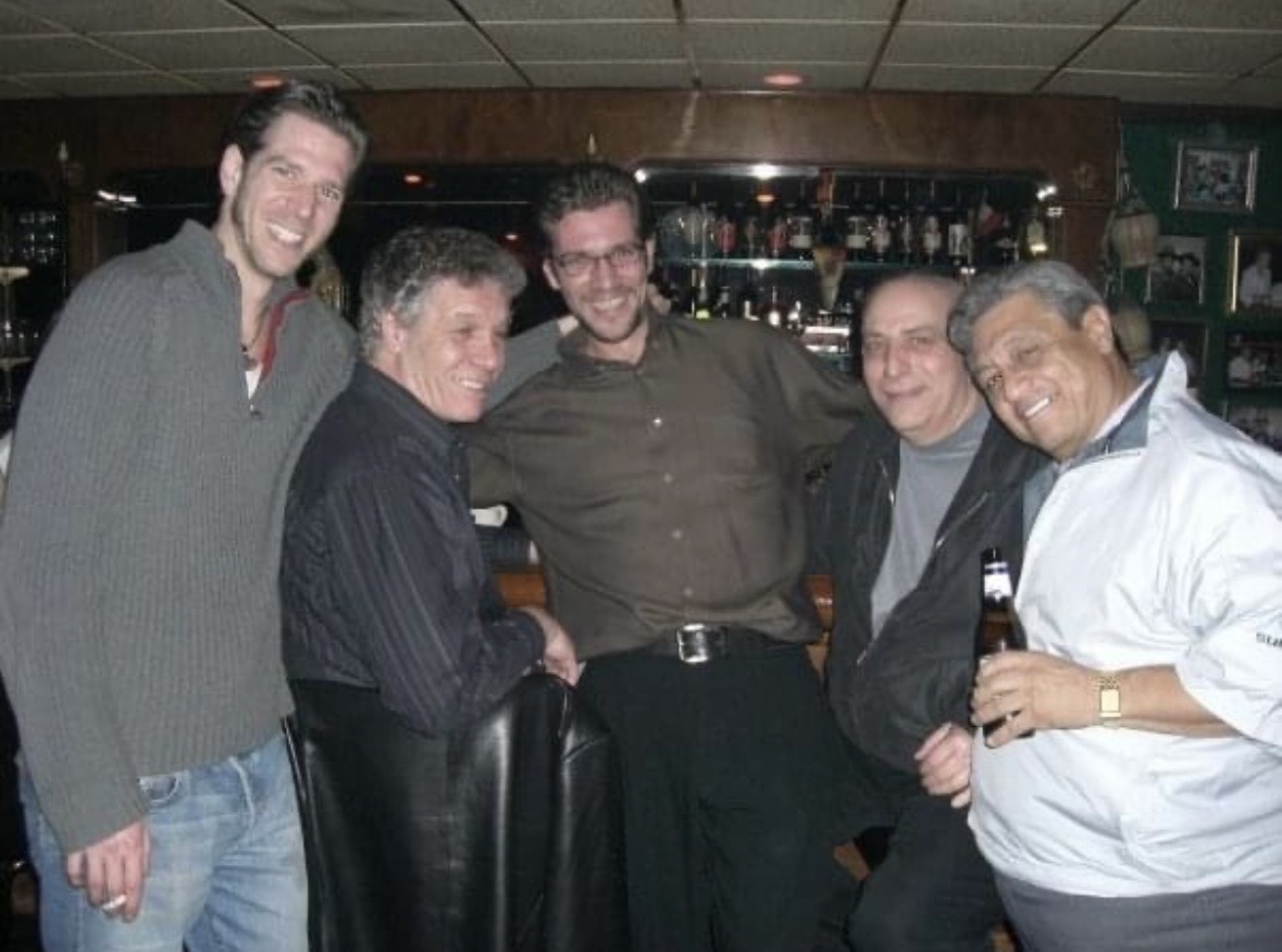 Five men standing together in a bar, smiling, with drinks in hand, in a dimly lit setting.