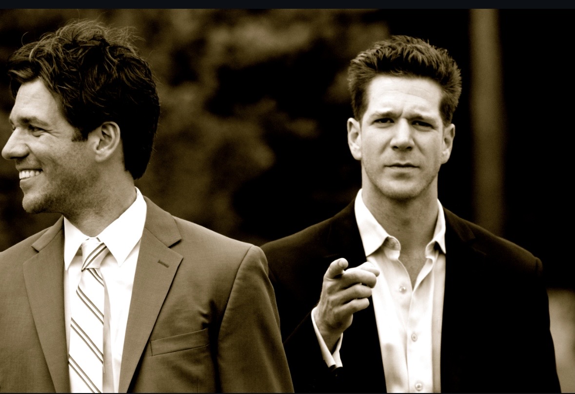 Two men in suits outdoors, one smiling and the other looking serious, with trees in the background.