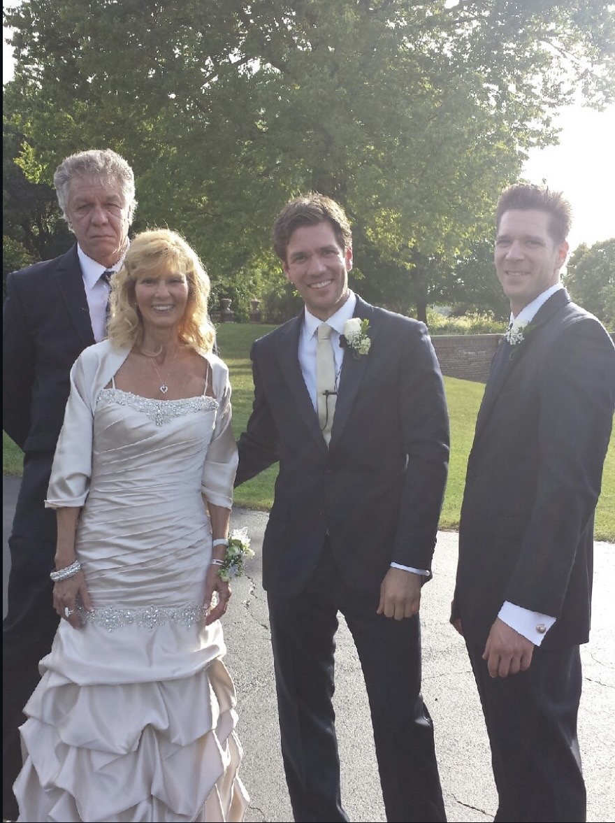 Four people dressed in formal attire standing outdoors on a sunny day, smiling, with trees and grass in the background.