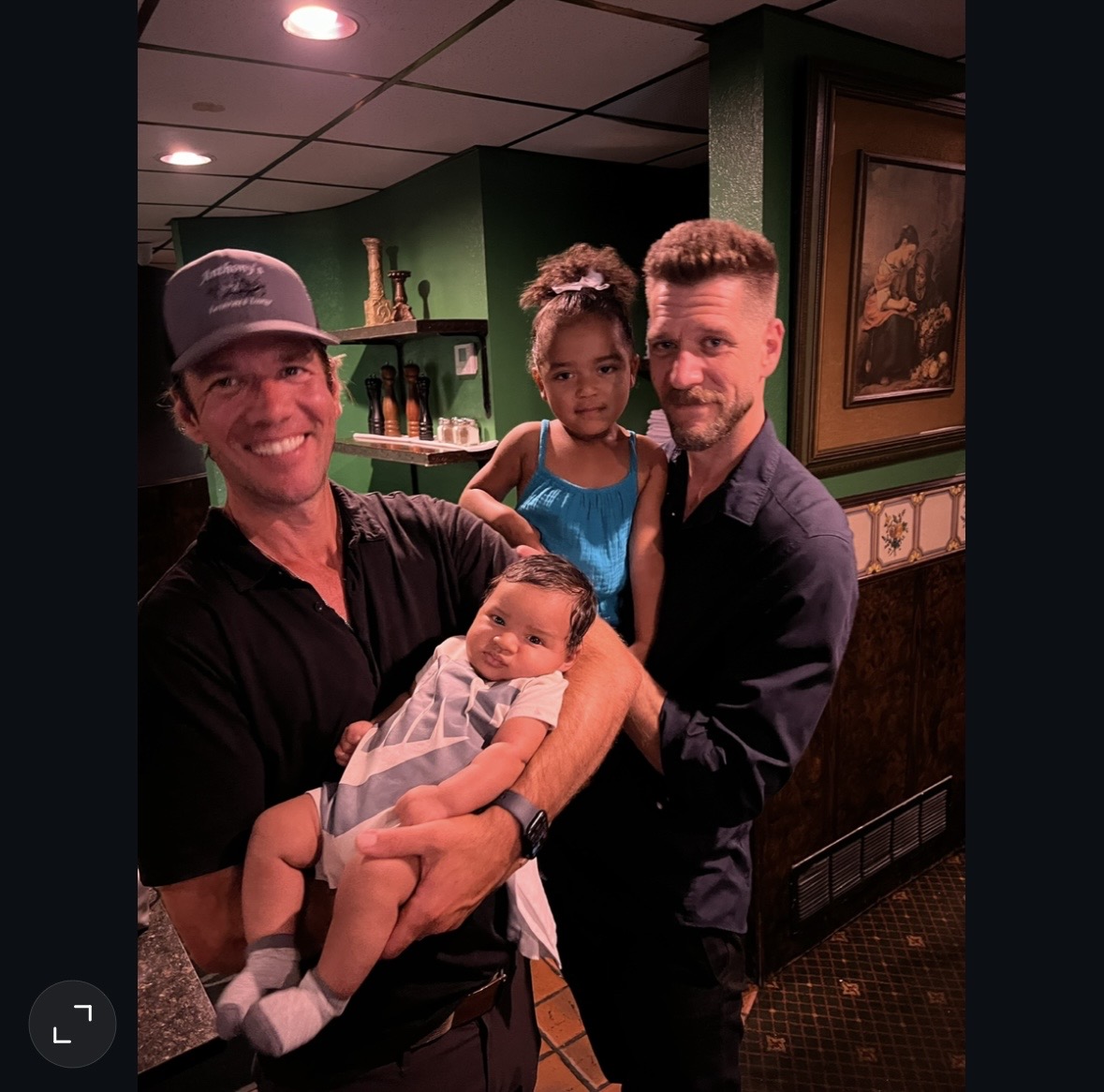 Two men, a girl, and a baby pose indoors with green walls and framed artwork, smiling at the camera.