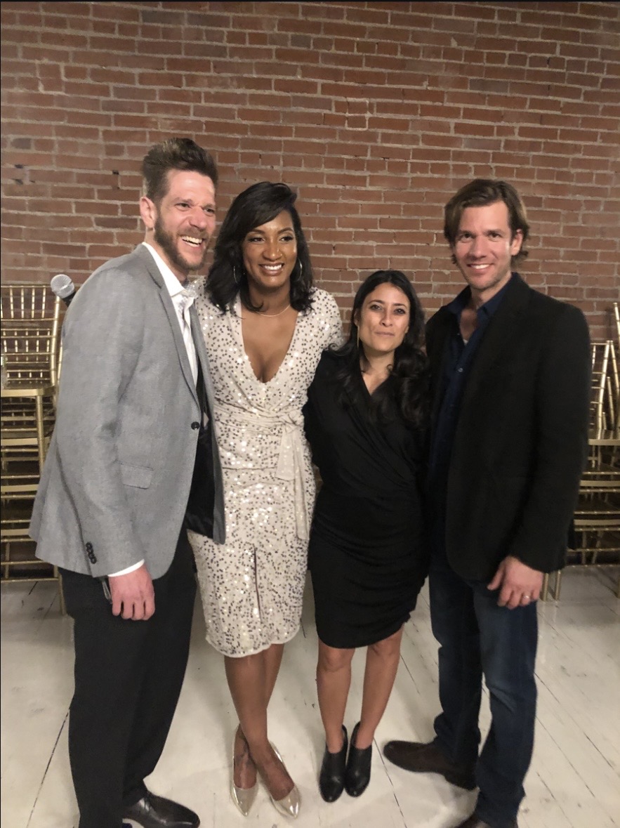 Four people standing together in front of a brick wall, smiling, dressed in formal attire.