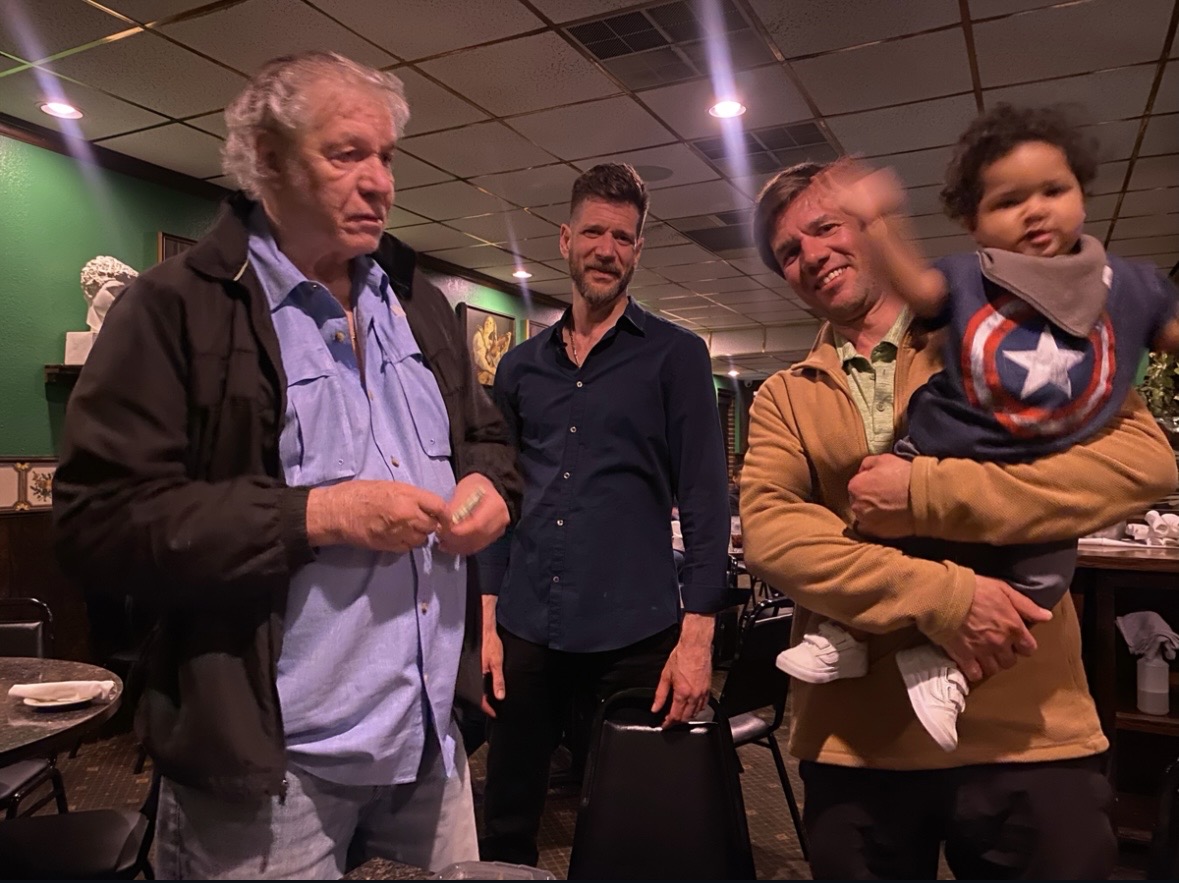 Four people, including a child, standing indoors in a room with a ceiling, some artwork, and a table, engaging in conversation.