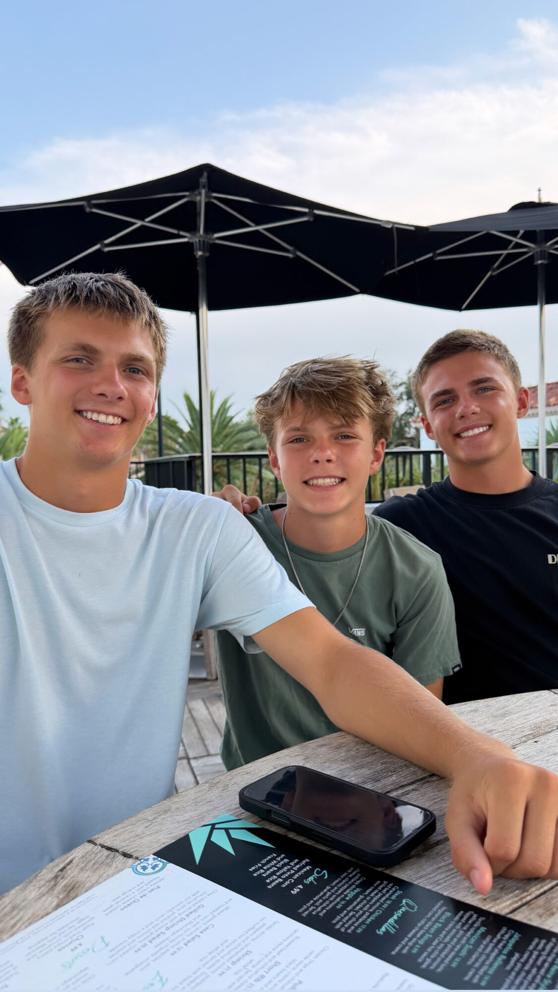 Three young men sitting outdoors at a table with umbrellas, smiling, with a menu and phone on the table.