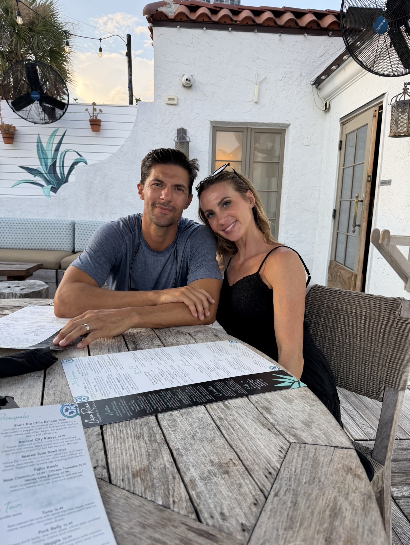 A man and woman sit at a table outdoors, smiling, with white walls, potted plants, and a door in the background.