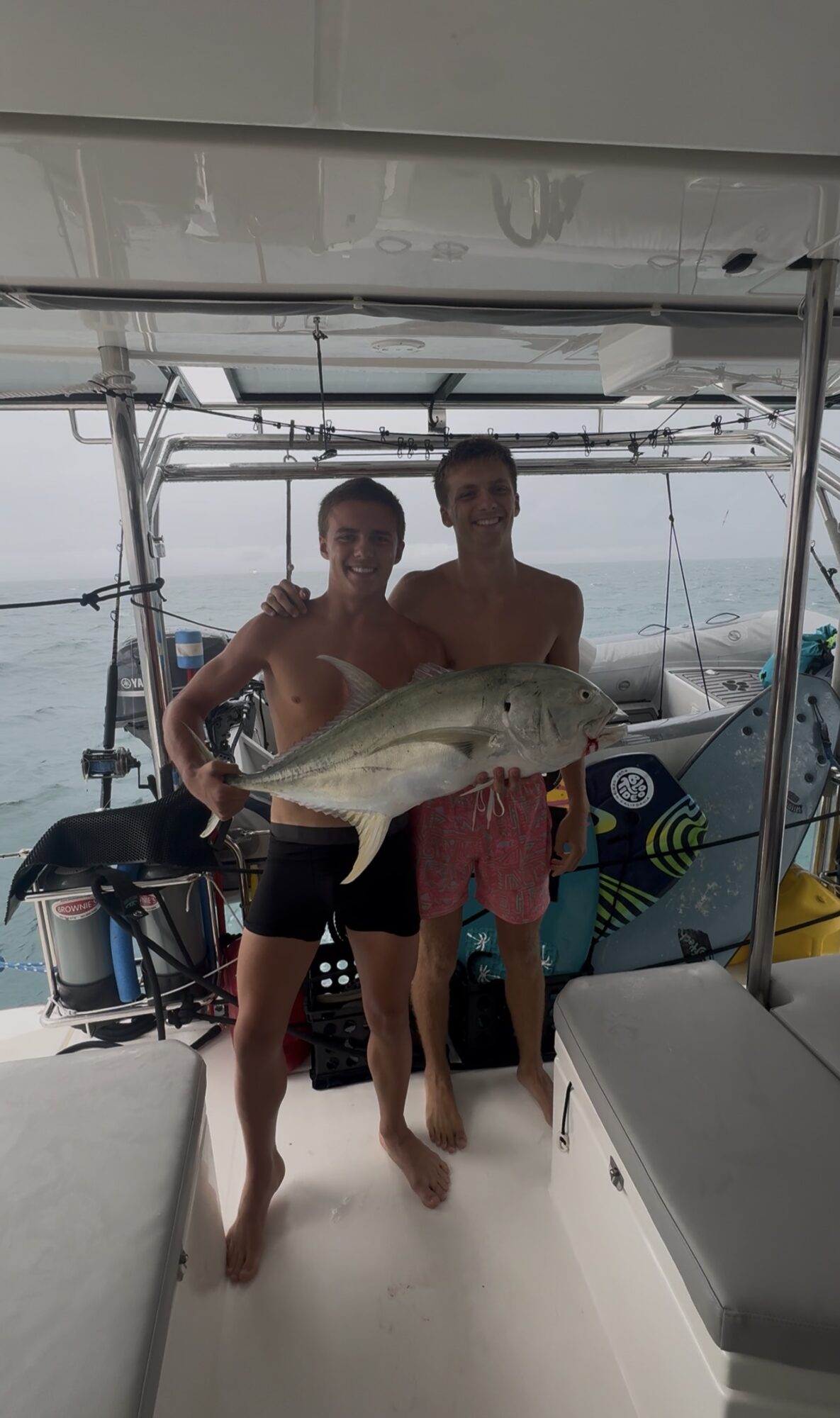 Two young men on a boat holding a large fish, with water and boat equipment in the background.
