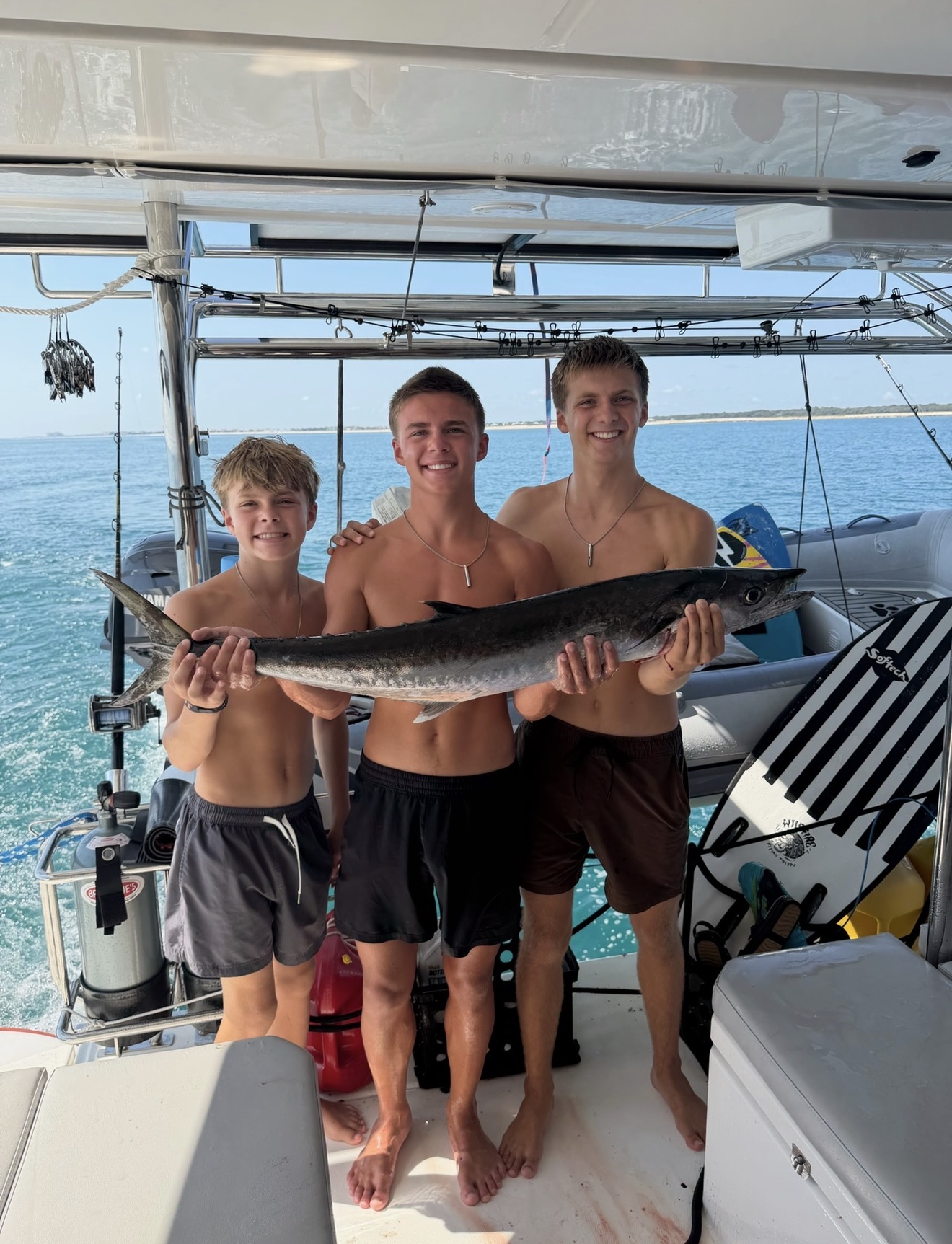 Three young men on a boat holding a large fish, with water and sky in the background.