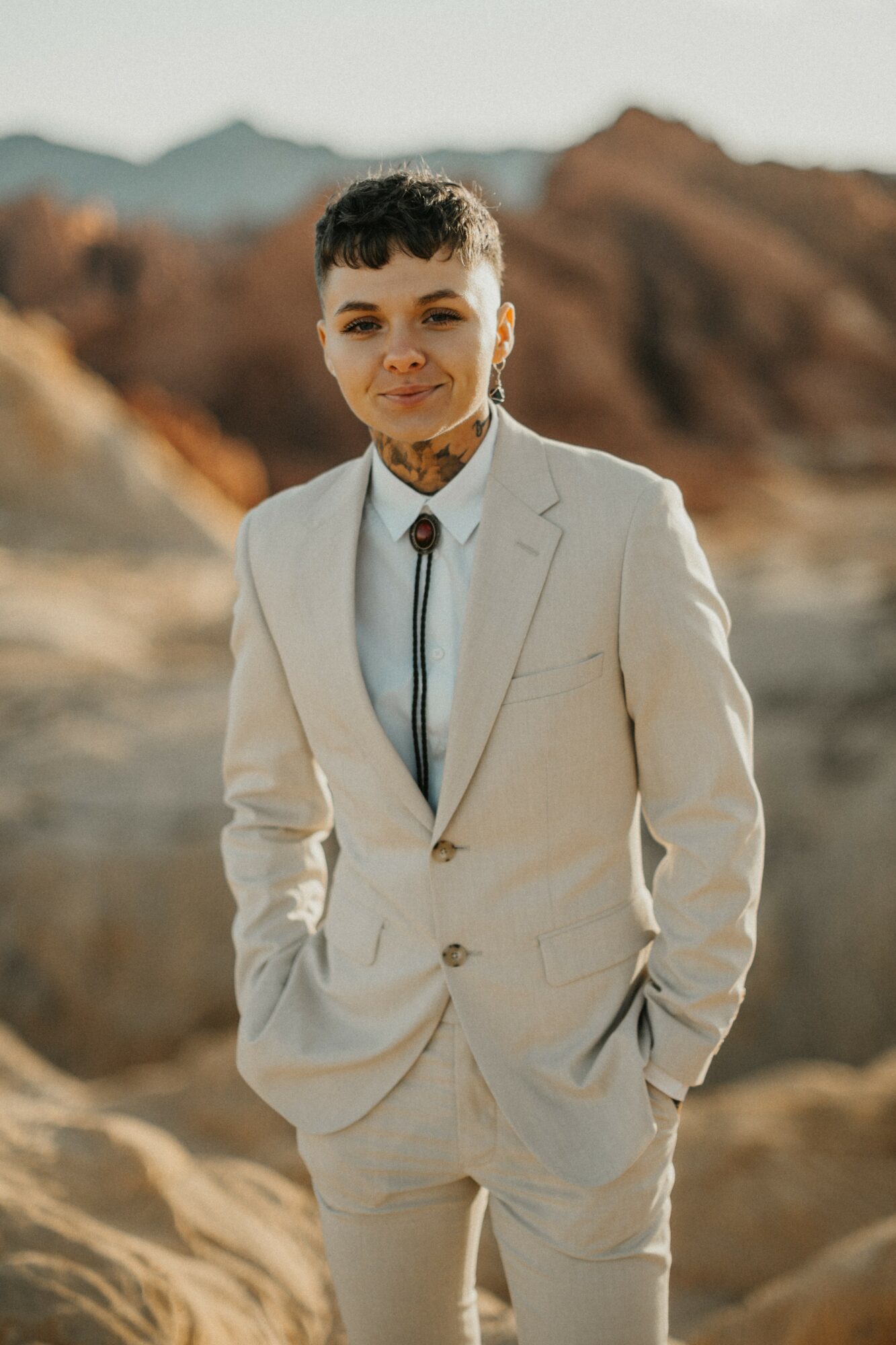 Young man in a light-colored suit outdoors with mountains in the background, smiling, hands in pockets.