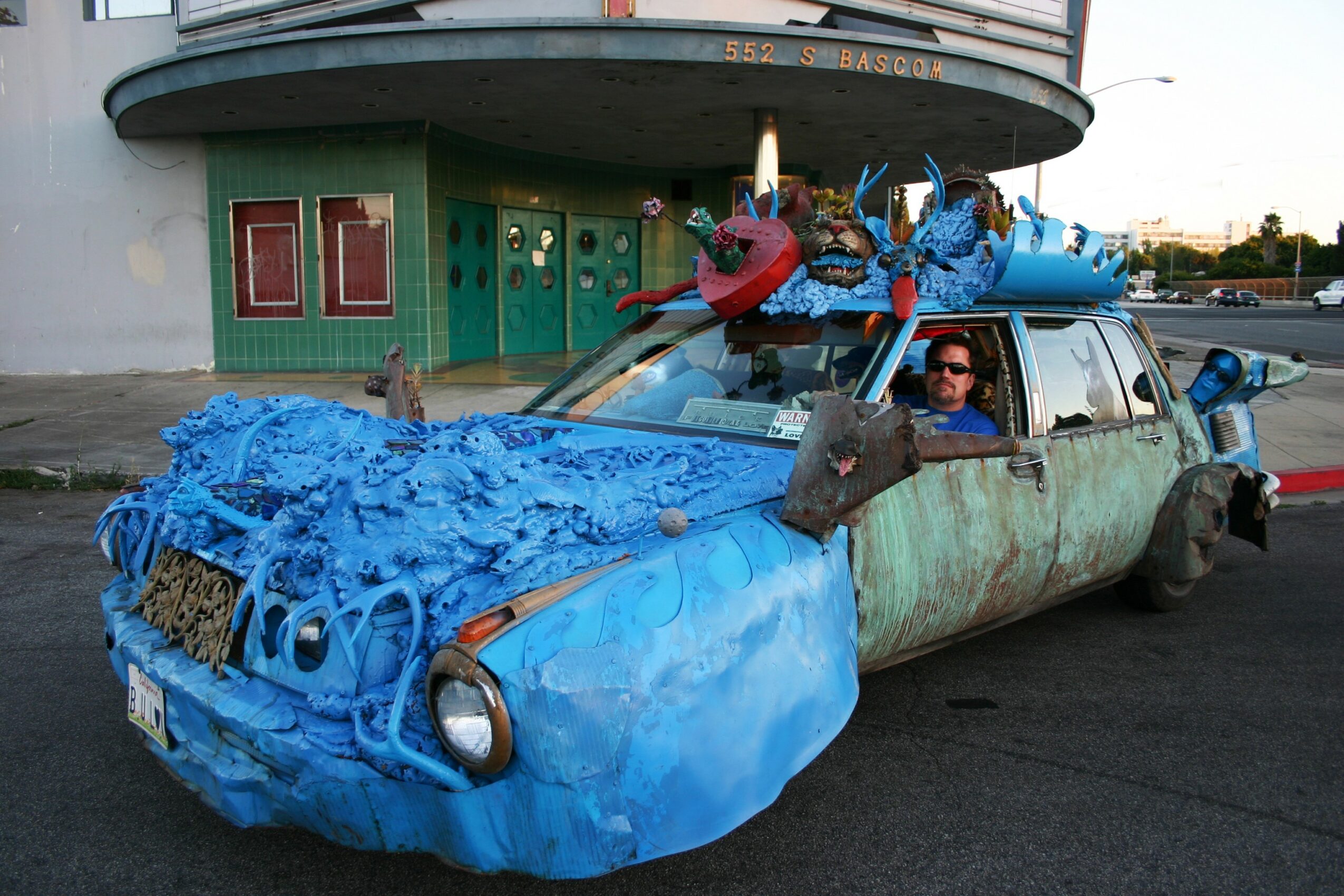 Car decorated with blue foam and various objects, with a person inside, parked in front of a building with green doors.