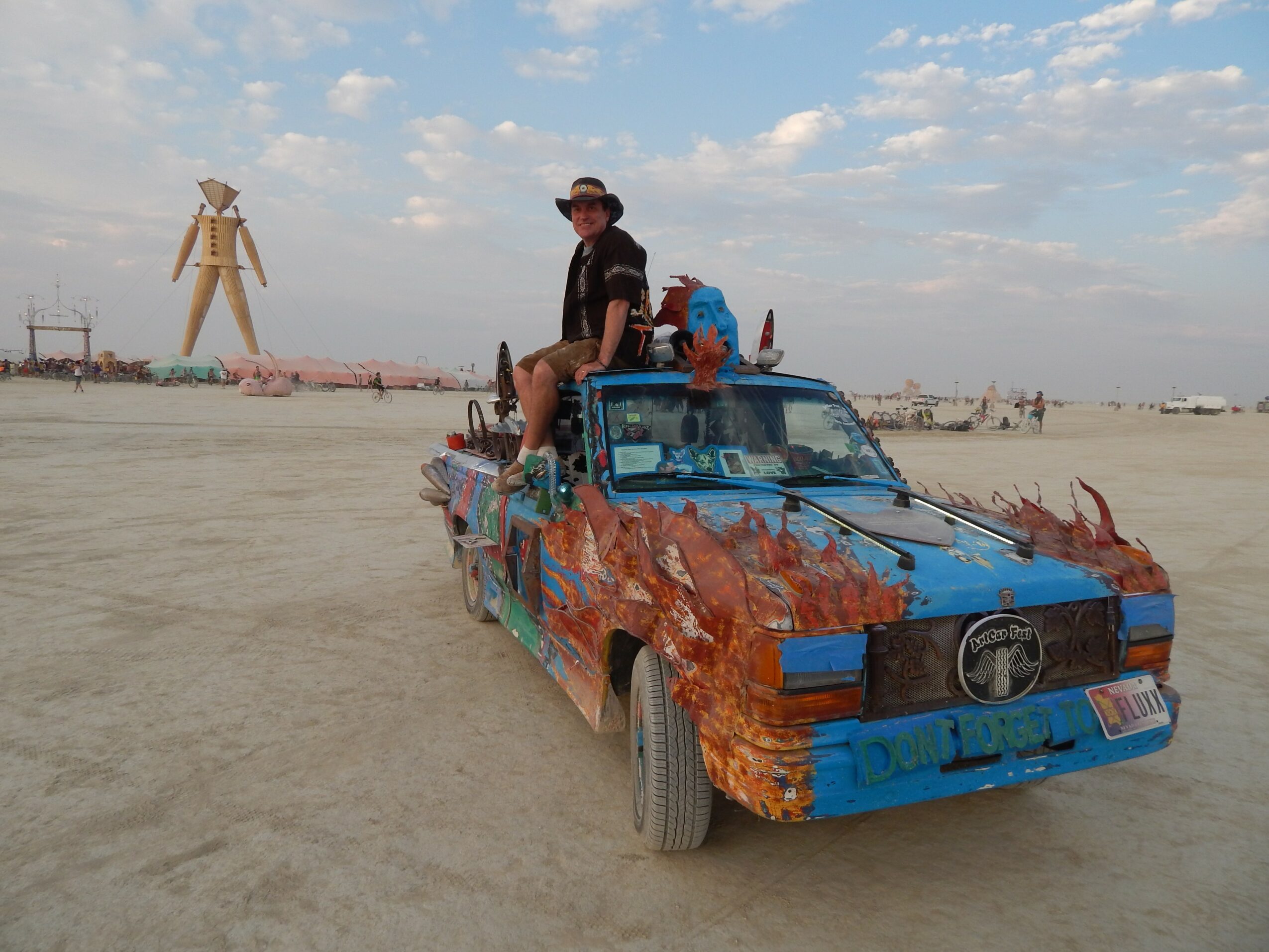 Person sitting on a decorated car with flames, in a desert landscape with a large wooden sculpture in the background.