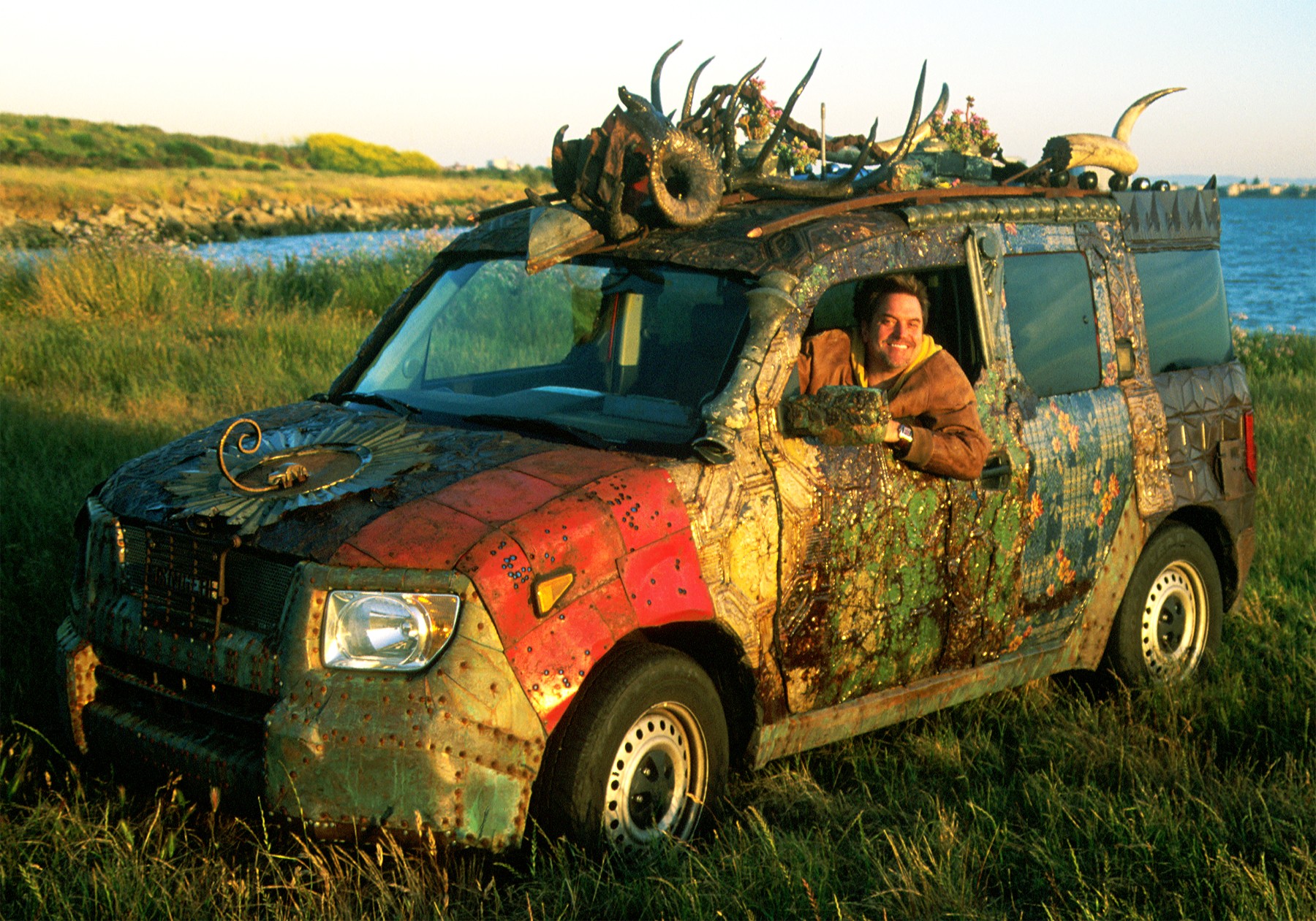 A camouflage-painted vehicle with a person inside, outdoors near a river, with debris on the roof and front, and grass around.