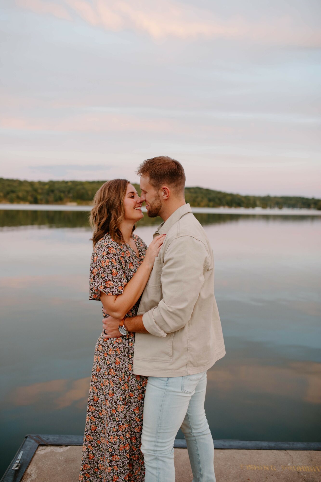 A couple stands close by a lake, gazing at each other, with a calm water and sky background.