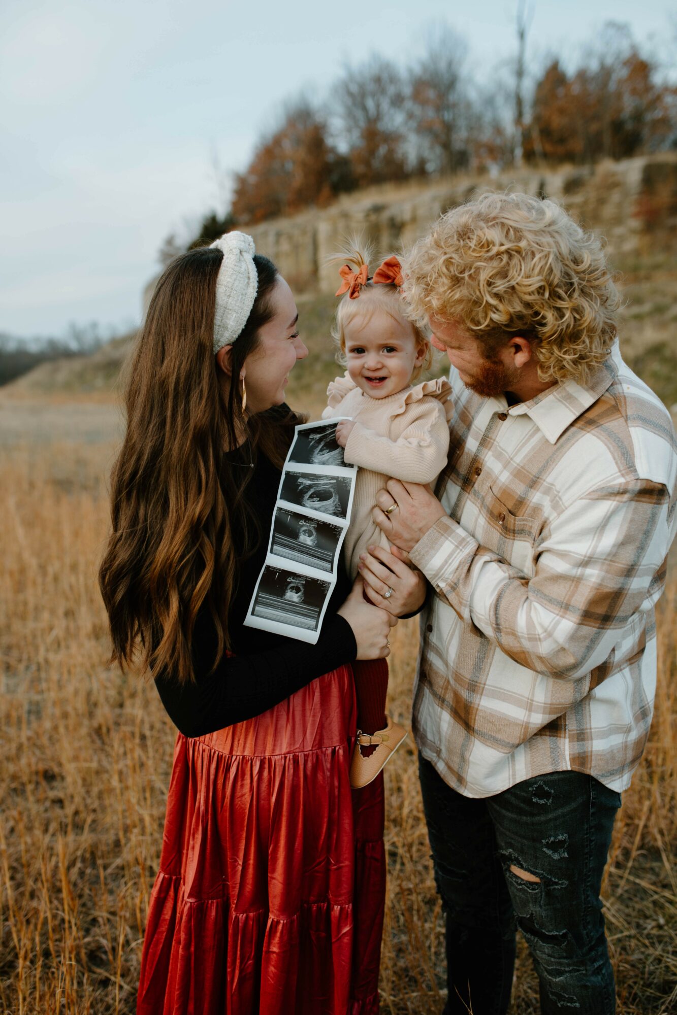 Family outdoors with a woman, man, and a smiling baby girl holding ultrasound images, in a grassy field with trees in background.
