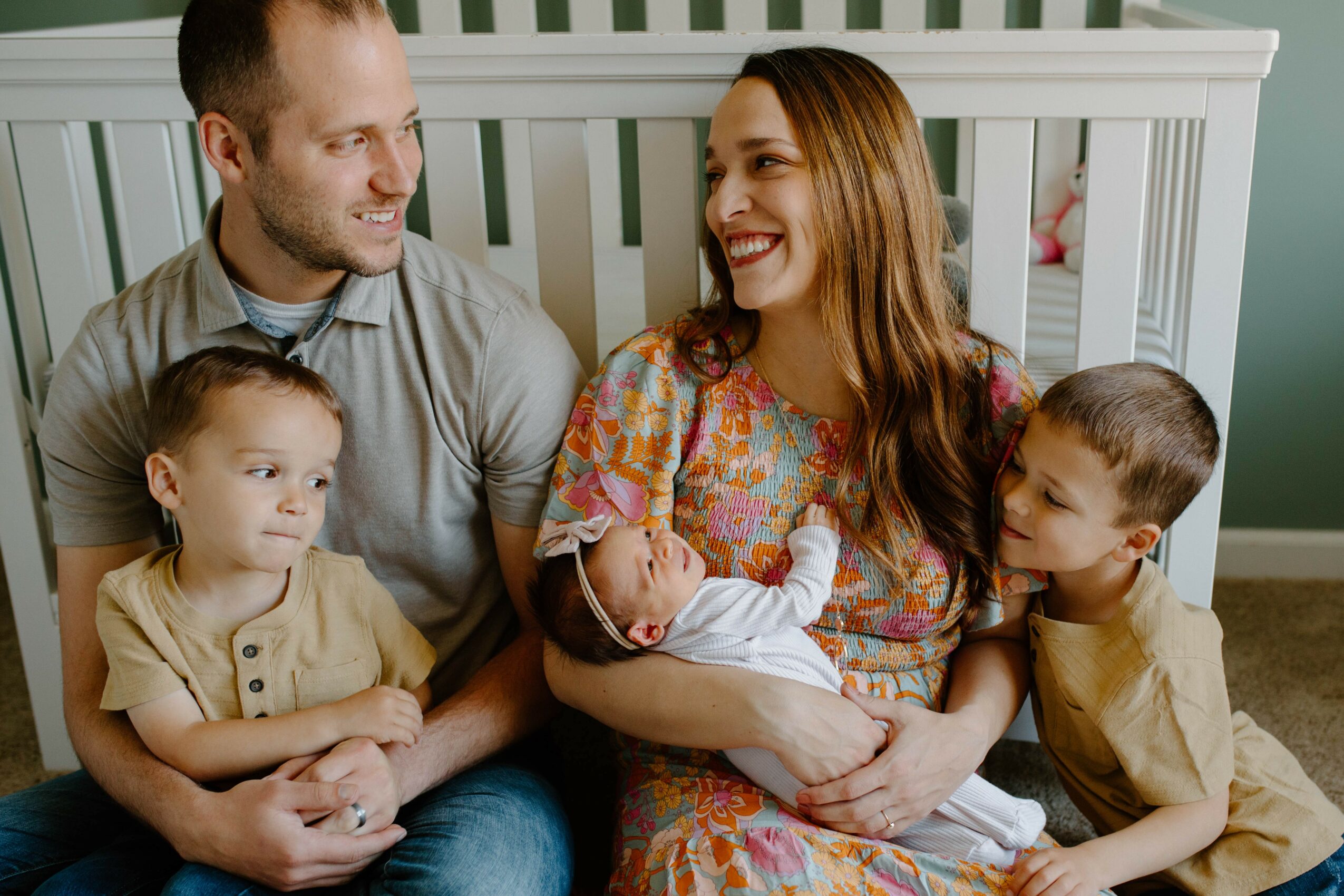 Family of four sitting together, smiling, with a baby in the mother's arms, in a room with a white crib.
