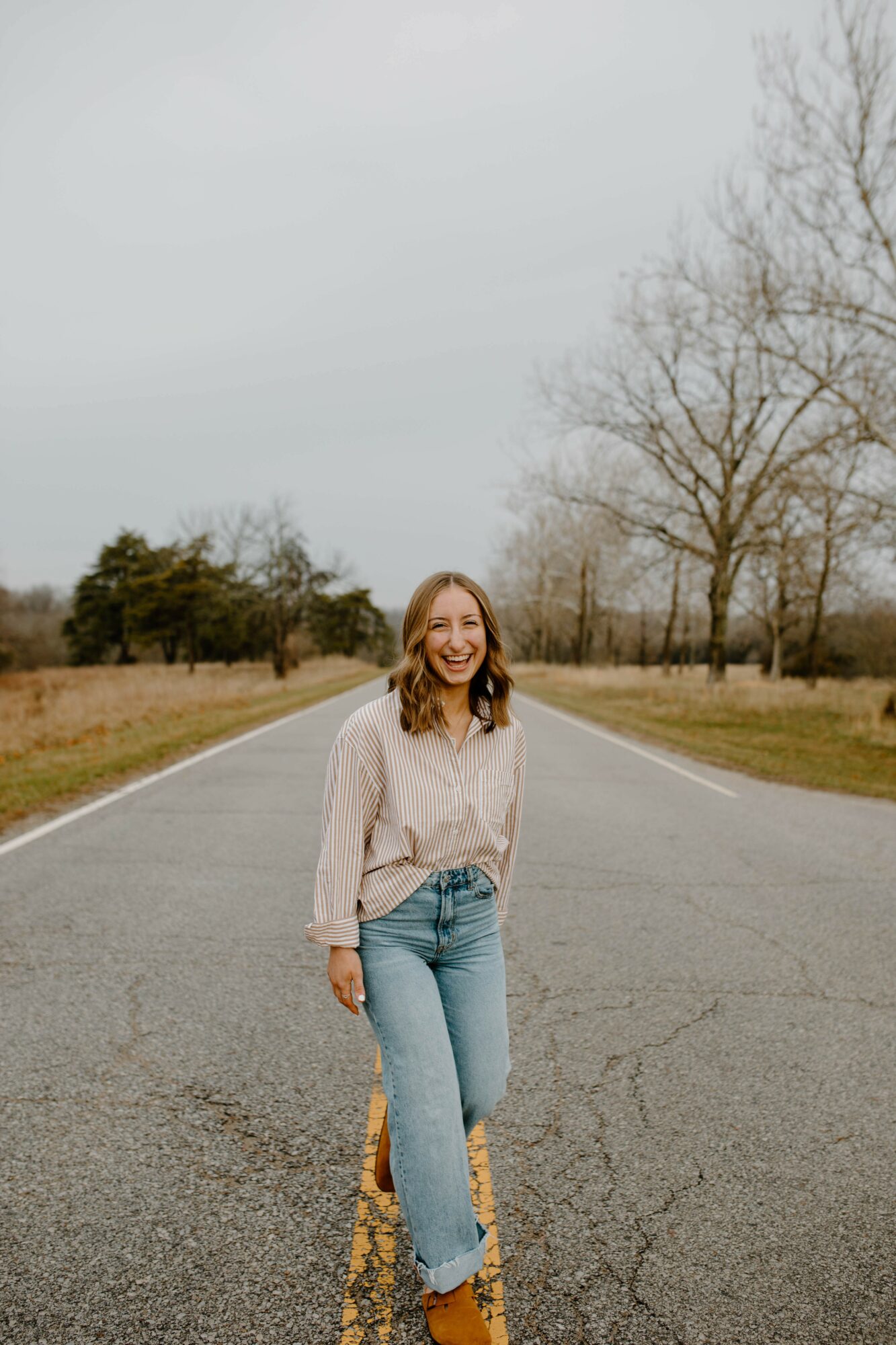 Young woman with long hair smiling, standing on an empty road in a rural area with trees and grass, overcast sky.