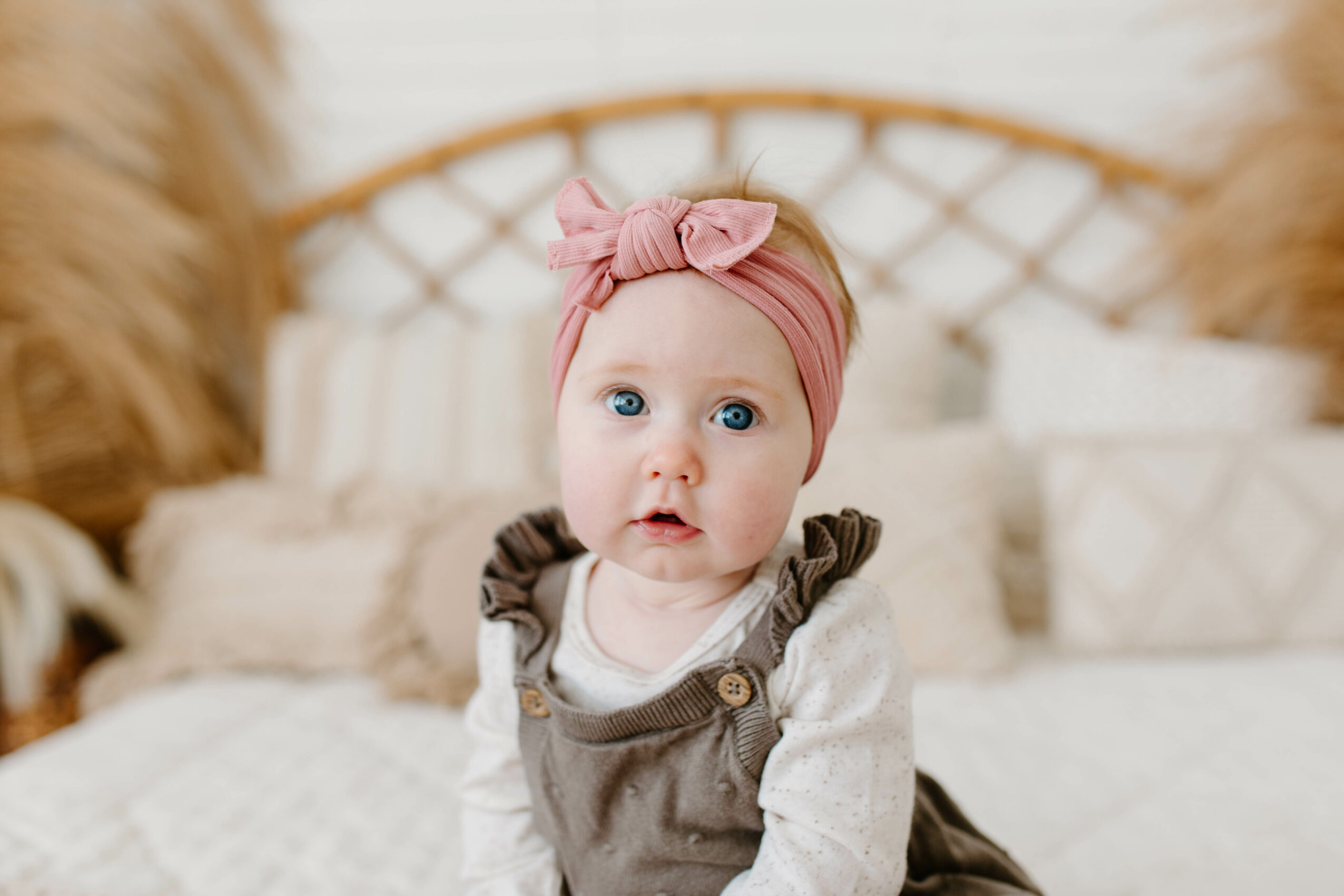 Baby girl with pink headband and bow, wearing overalls, sitting on bed with pillows, looking at camera.