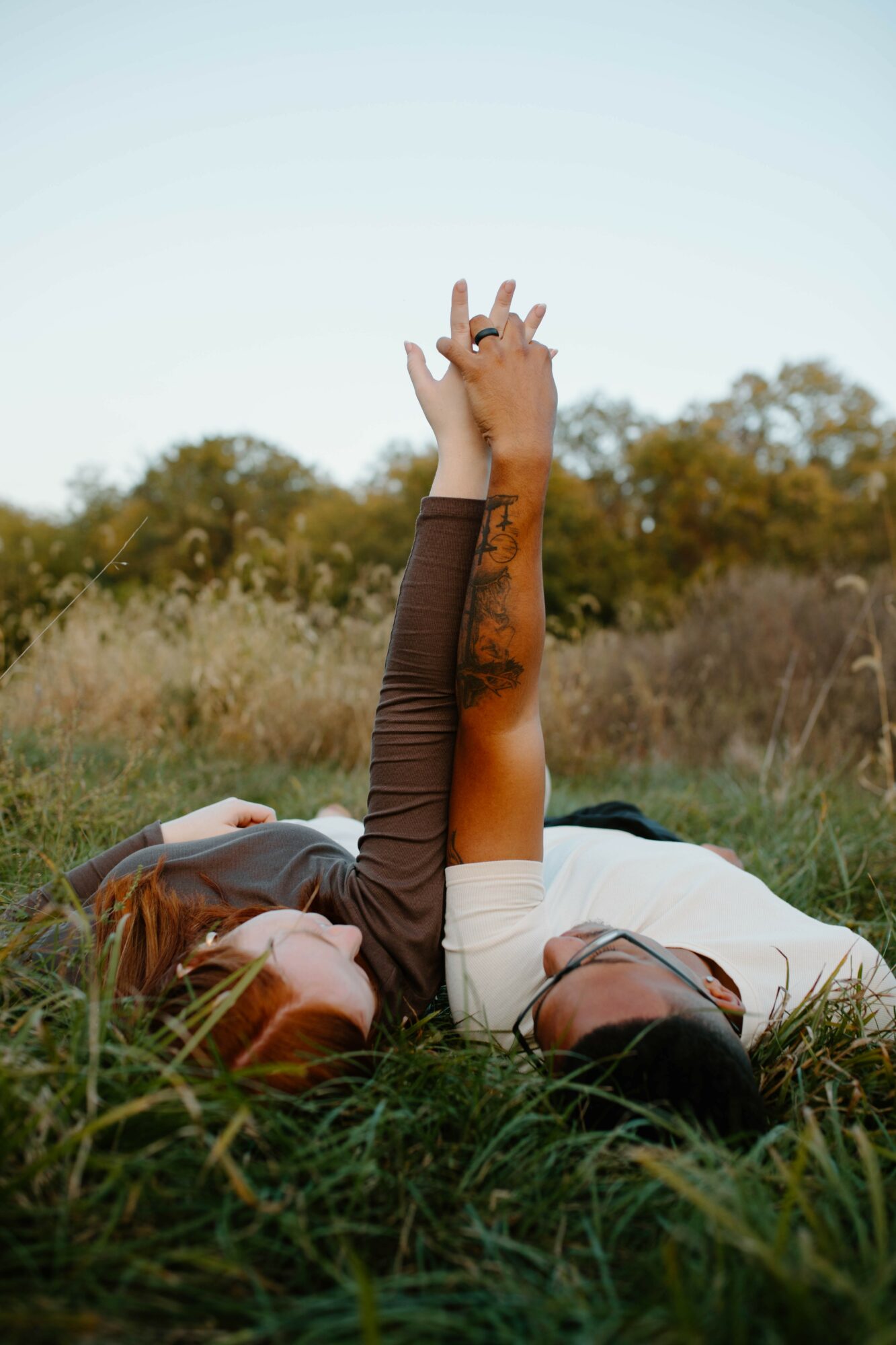 Two people lying on grass, holding hands with one arm raised, outdoors with trees in the background.