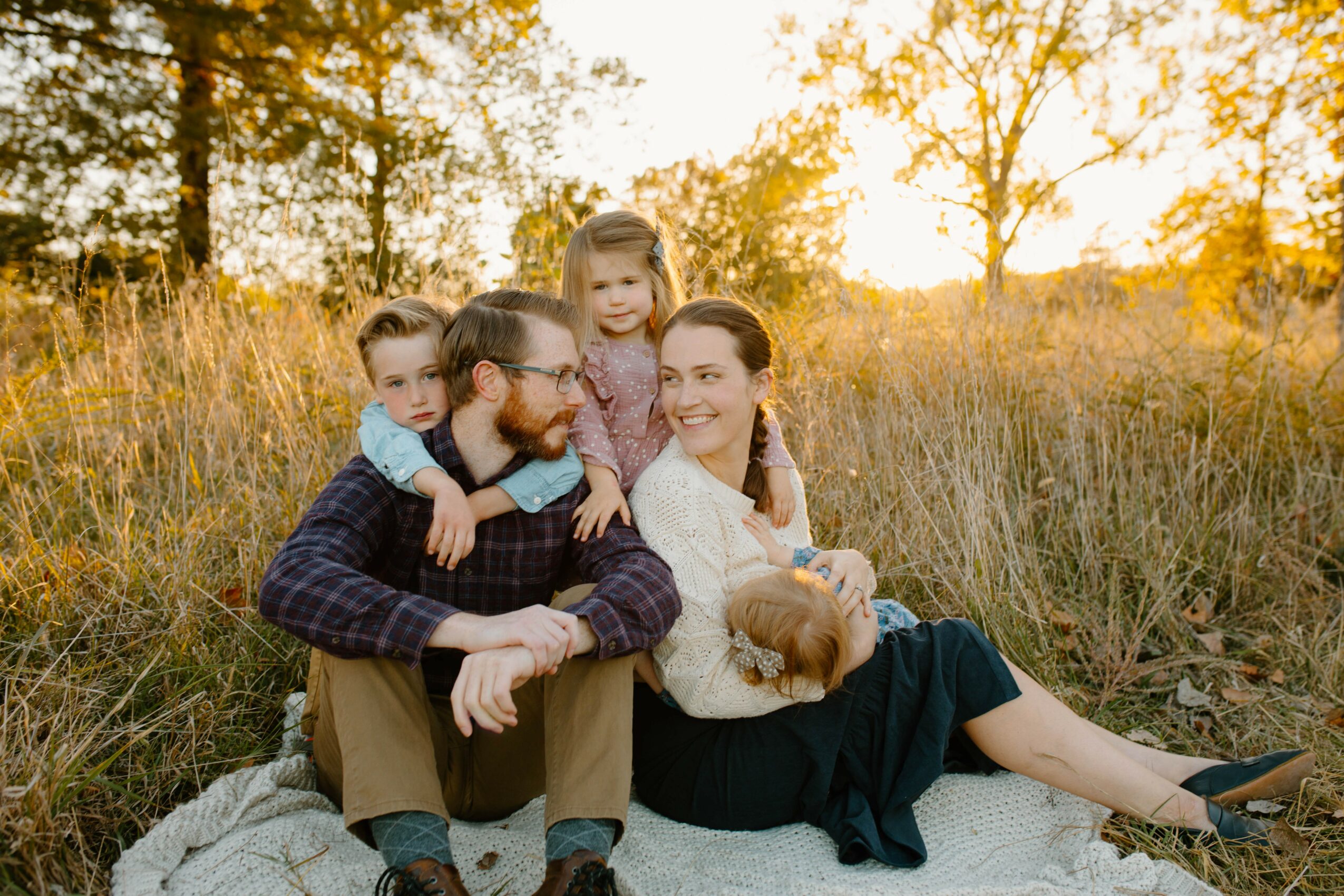 Family of four sitting on a blanket outdoors during sunset, smiling and enjoying each other's company.