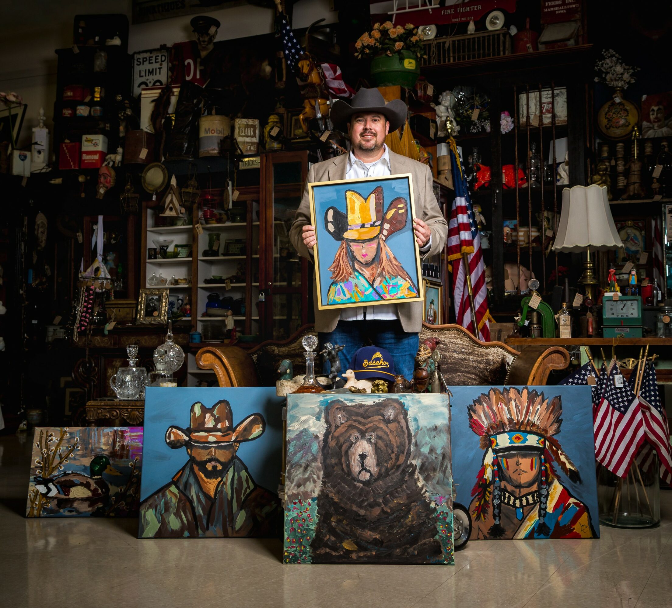 Person holding a colorful portrait of a bear, surrounded by paintings of a bear, a cowboy, and a Native American, in an antique shop.