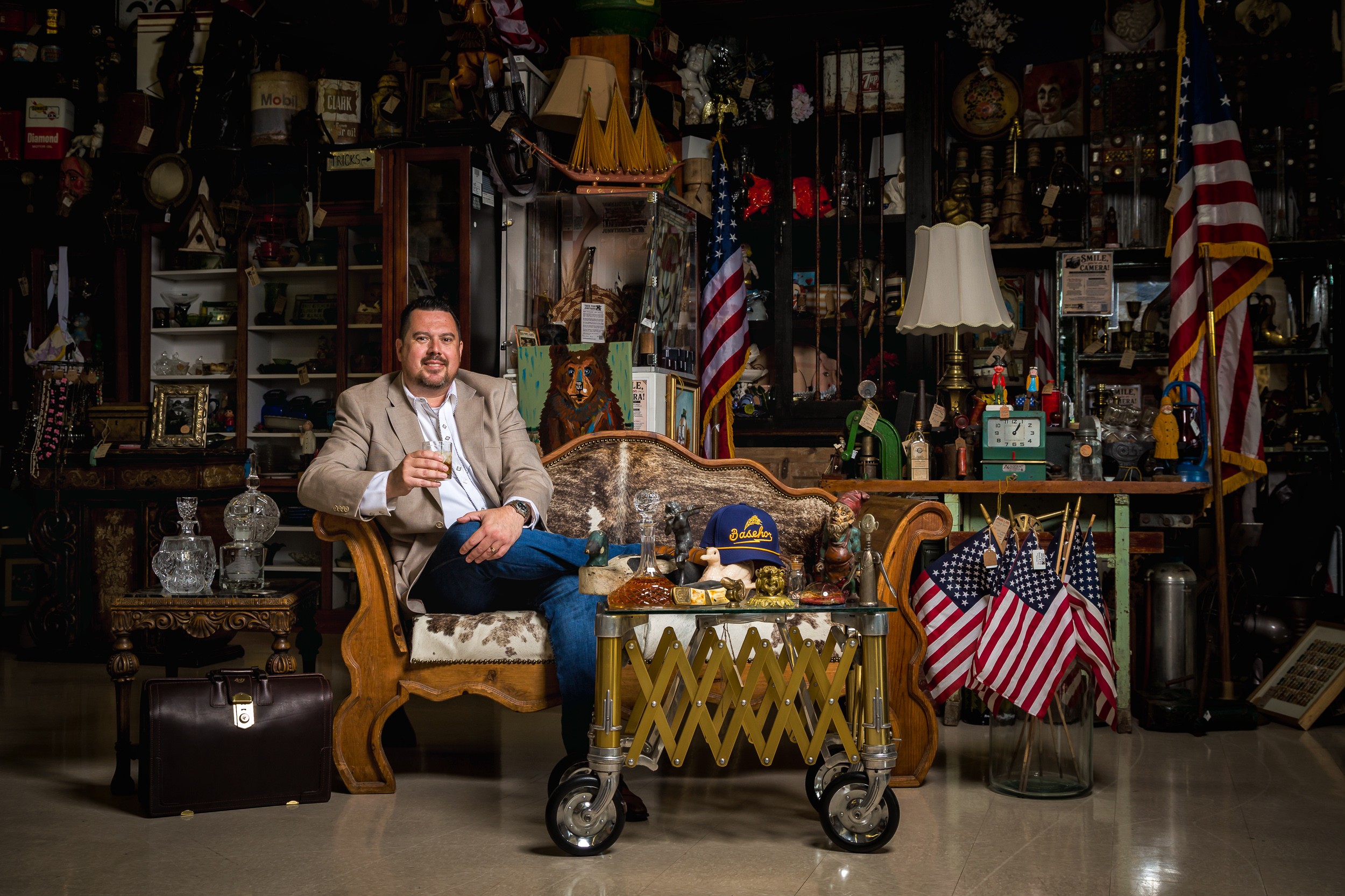 Man sitting on a vintage sofa in a room filled with American flags, lamps, and various decorative items.