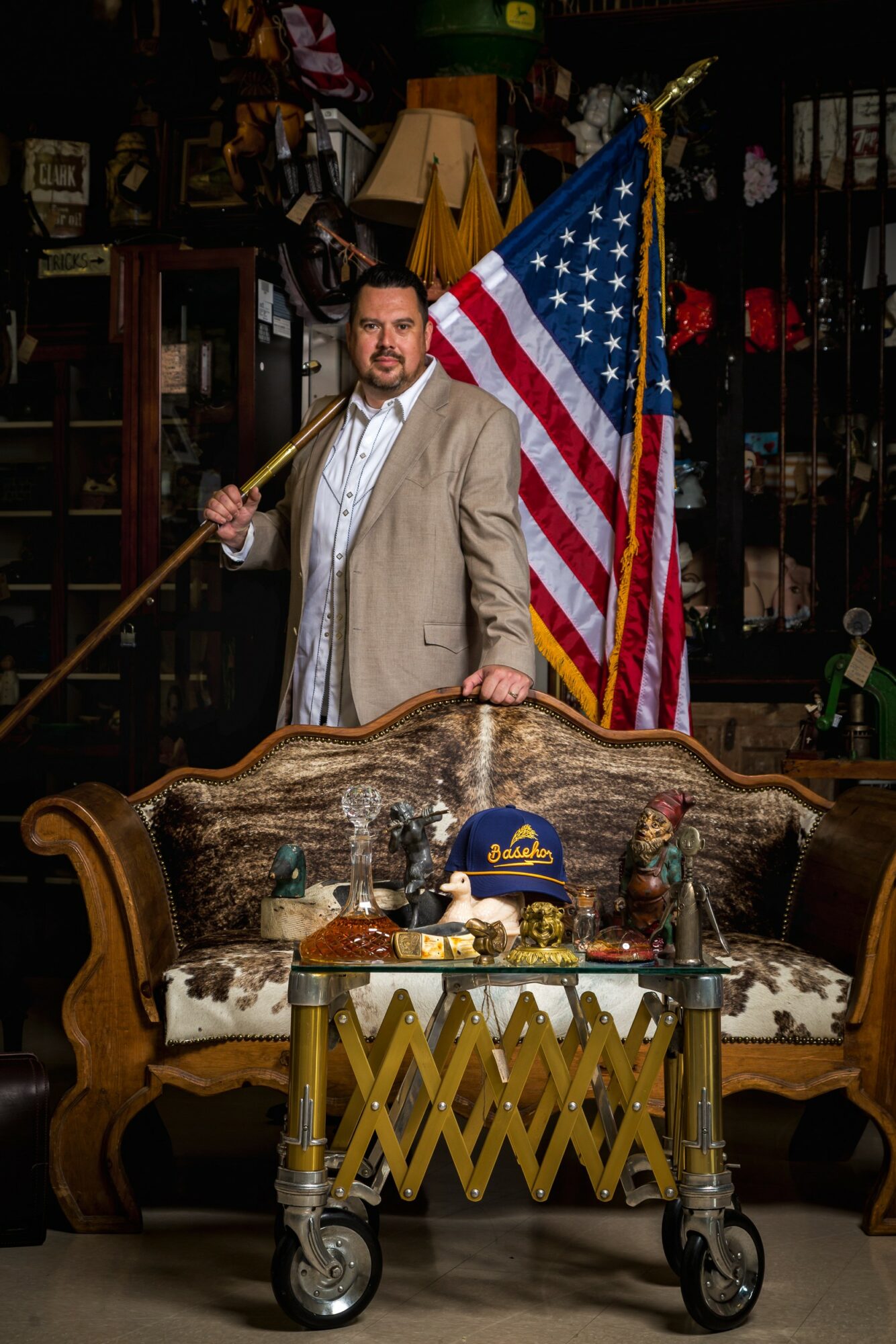 Man holding an American flag in a room with vintage furniture and decor, standing behind a wooden sofa and a cart.