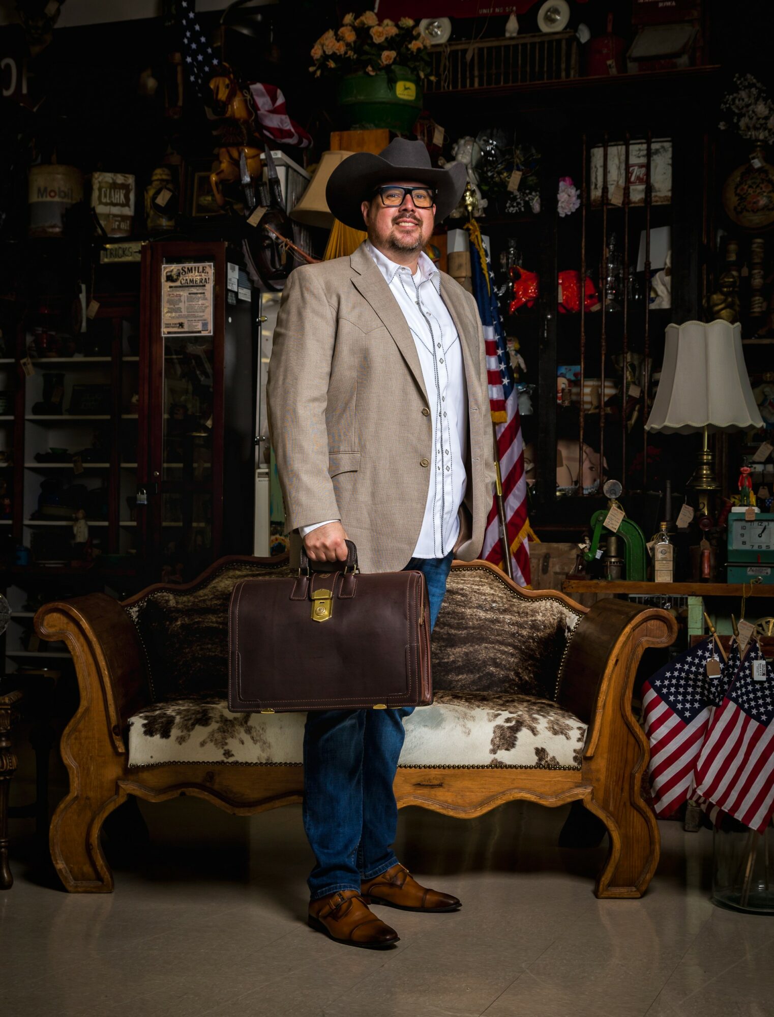 Man in cowboy hat and glasses holding a briefcase, standing in a vintage store with furniture and decorations.