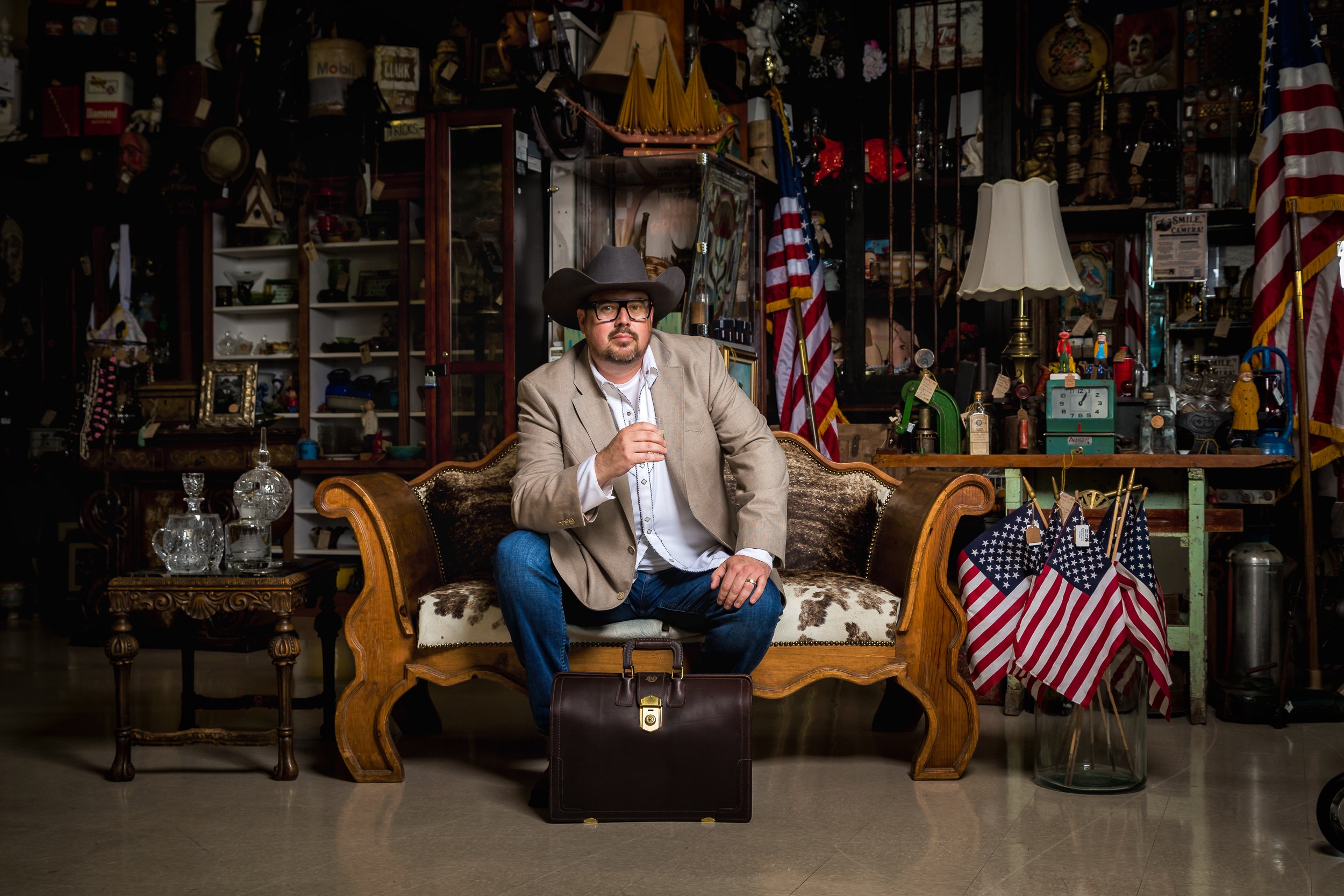 Man sitting on a vintage sofa in a store with American flags and patriotic decorations, holding a drink, with a suitcase in front.