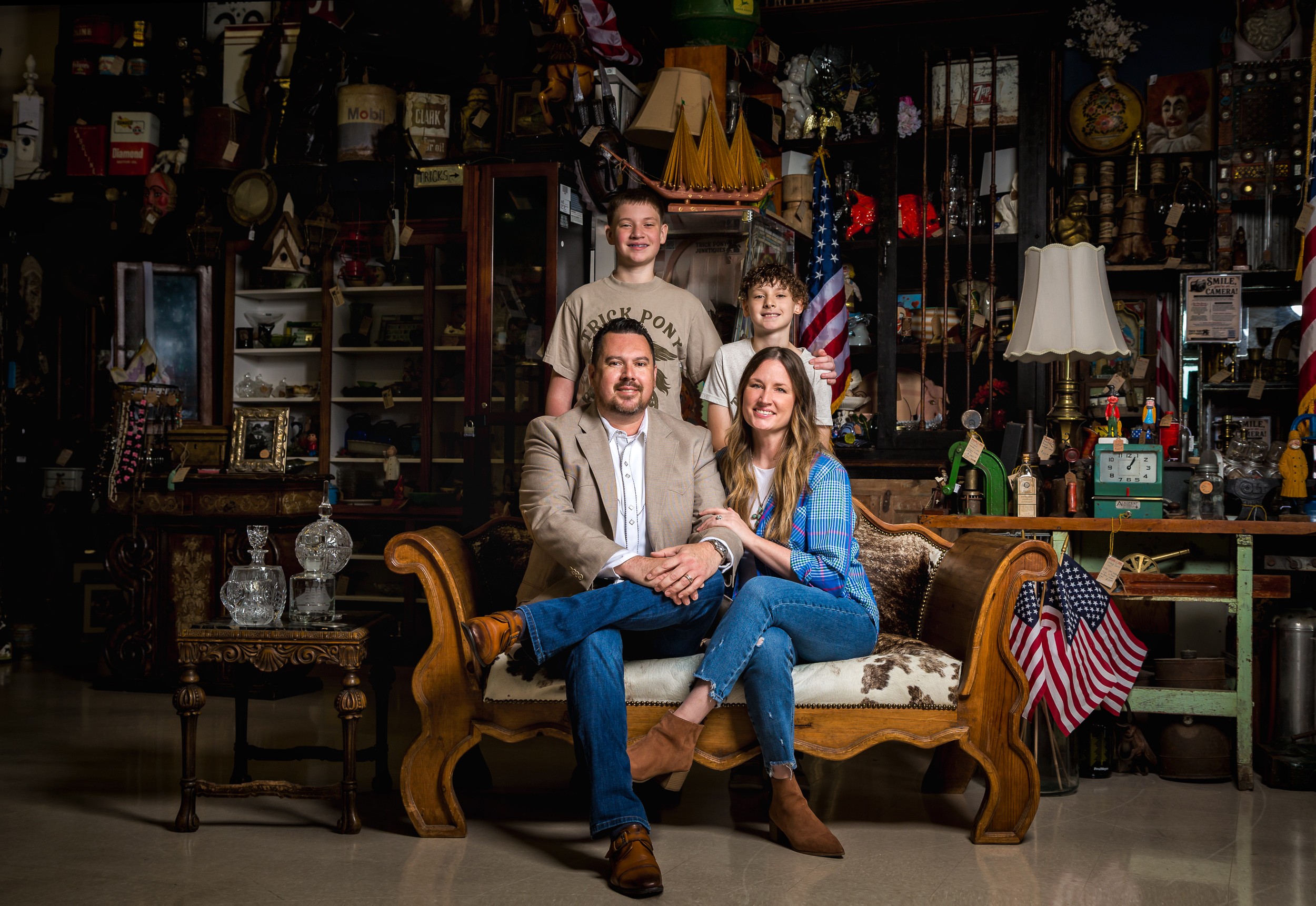 Family of four sitting on a vintage sofa in a cluttered room filled with collectibles and decorations.