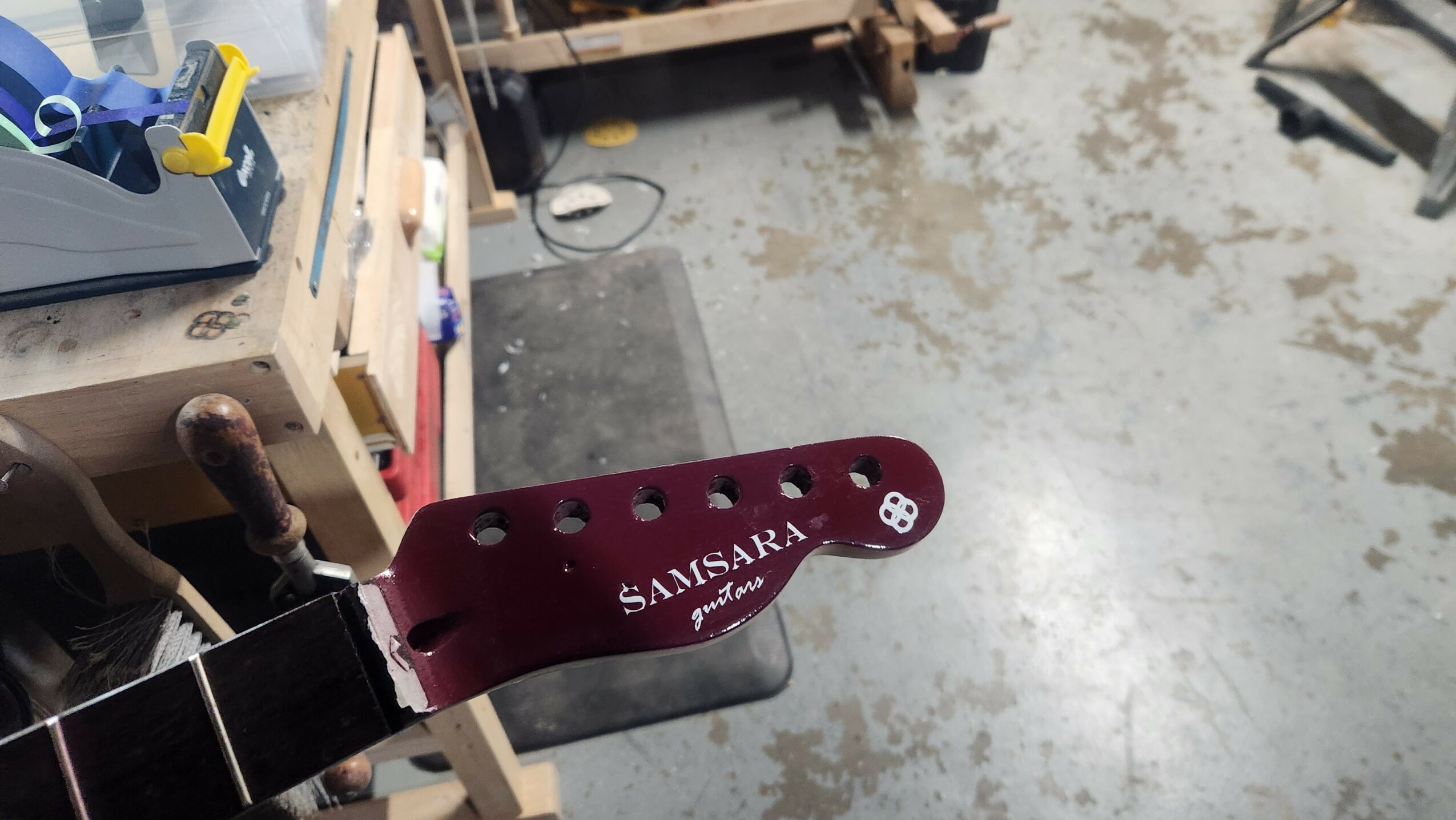 Close-up of a guitar headstock with tuning pegs and a maroon logo plate reading 'SAMSAARA' in a workshop.