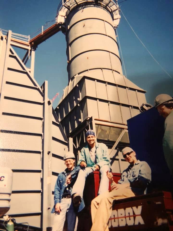 Group of four people sitting and standing near a large industrial structure with a clear sky background.