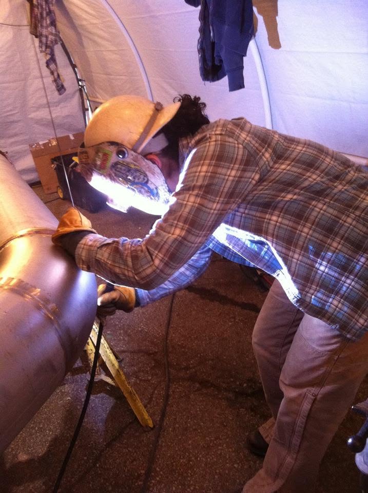 Person welding inside a tent, wearing protective gear, working on a large metallic object, with tools and equipment nearby.