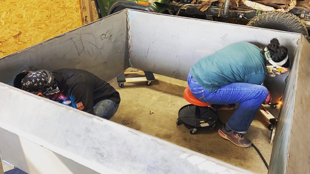 Two people are kneeling inside a small, enclosed space, working on something on the floor. One wears a black jacket, the other a blue jacket.