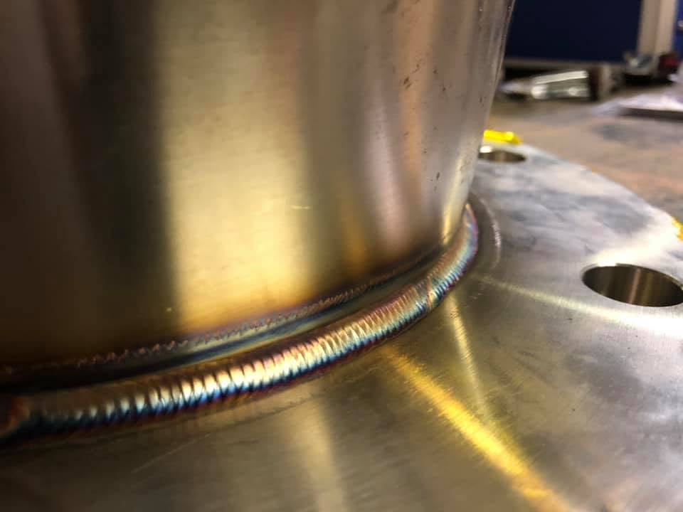 Close-up of a metal weld on a circular metal piece, showing rainbow-colored heat discoloration and a smooth weld bead.