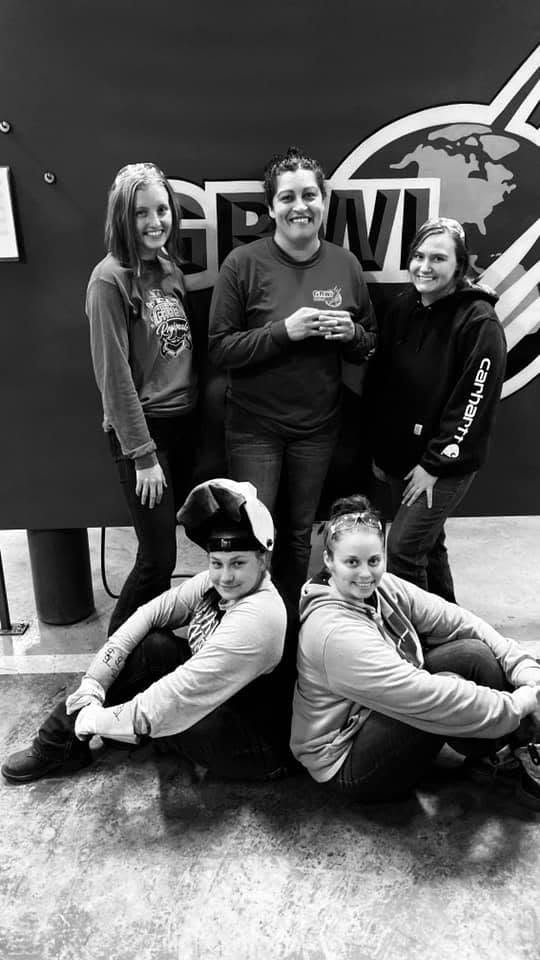 Four people, three standing and two sitting on the floor, in front of a wall with a logo, smiling.